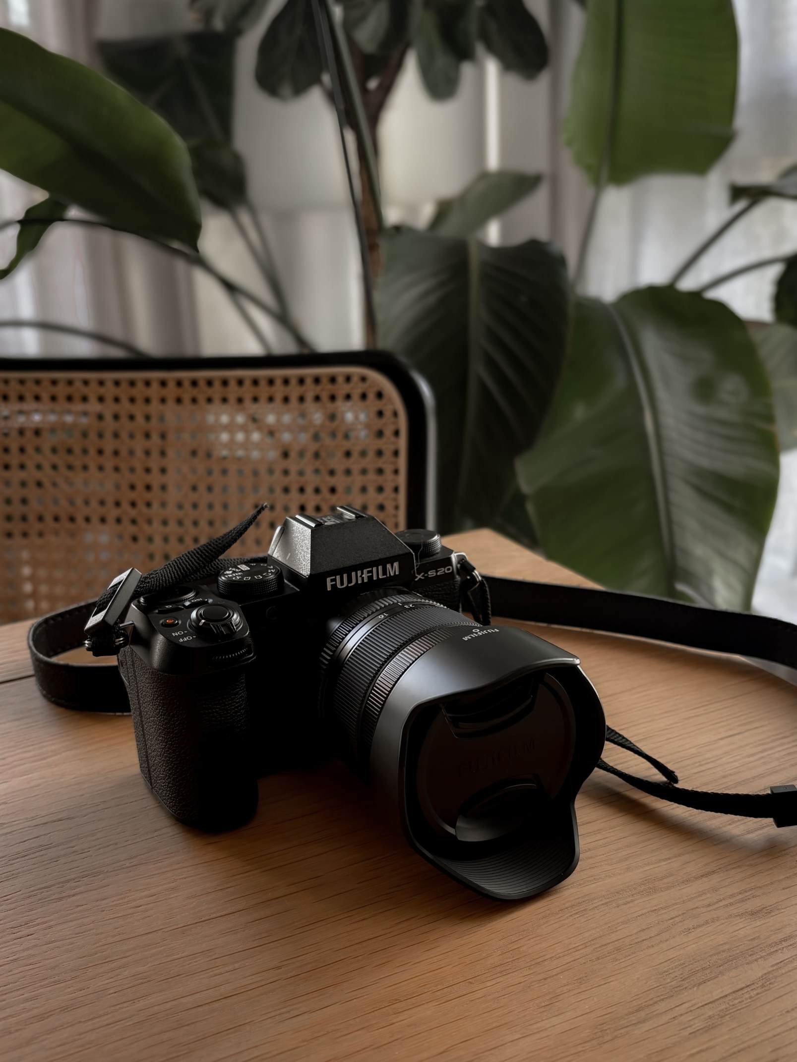 Un appareil photo Fujifilm X-S20 posé sur une table en bois. En arrière-plan, un fauteuil en osier et plusieurs grandes feuilles de plantes vertes.
