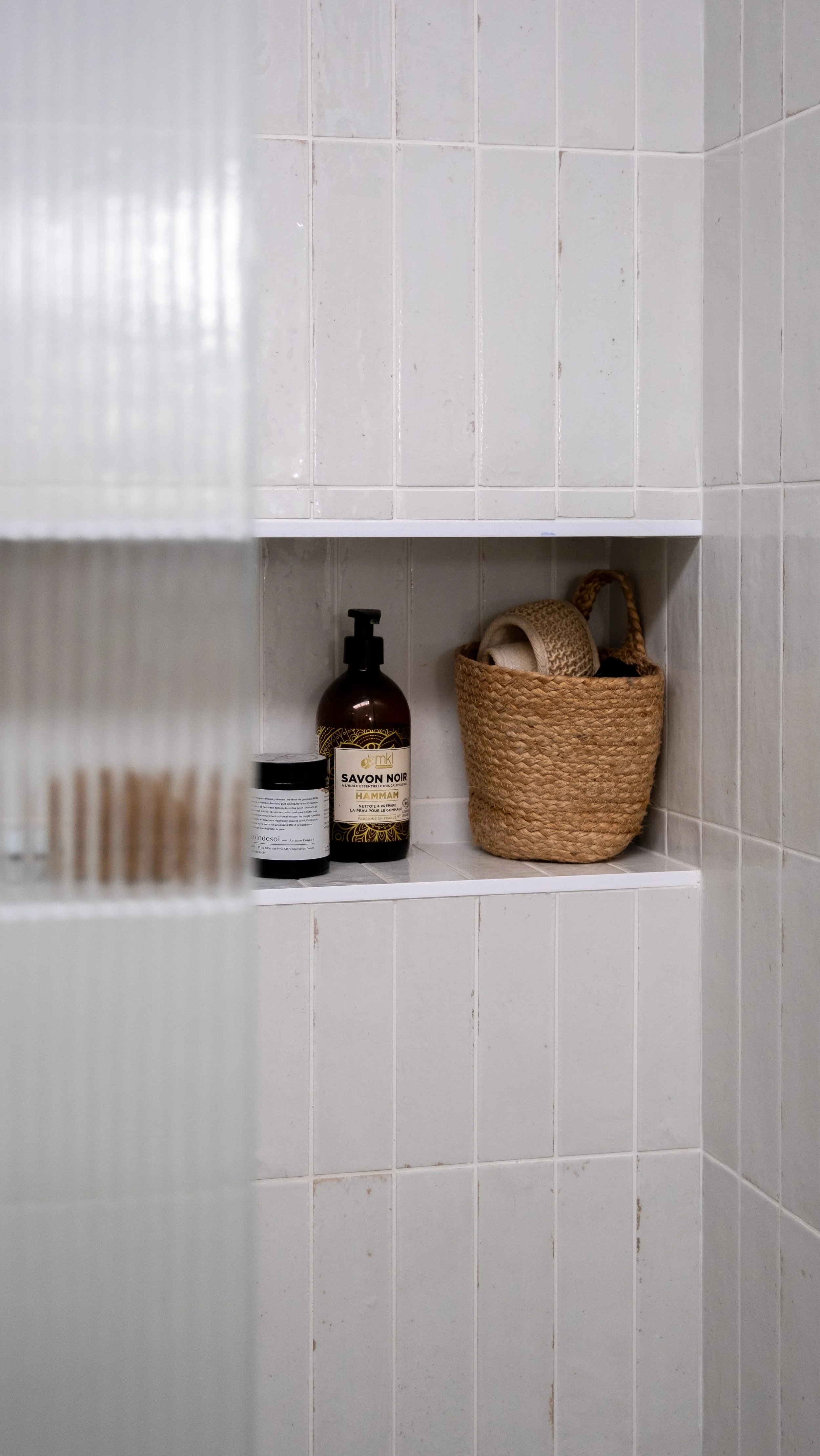 Vue partielle d'une niche dans une douche avec un flacon de savon noir, un petit pot et un panier en rotin contenant des accessoires de bain.