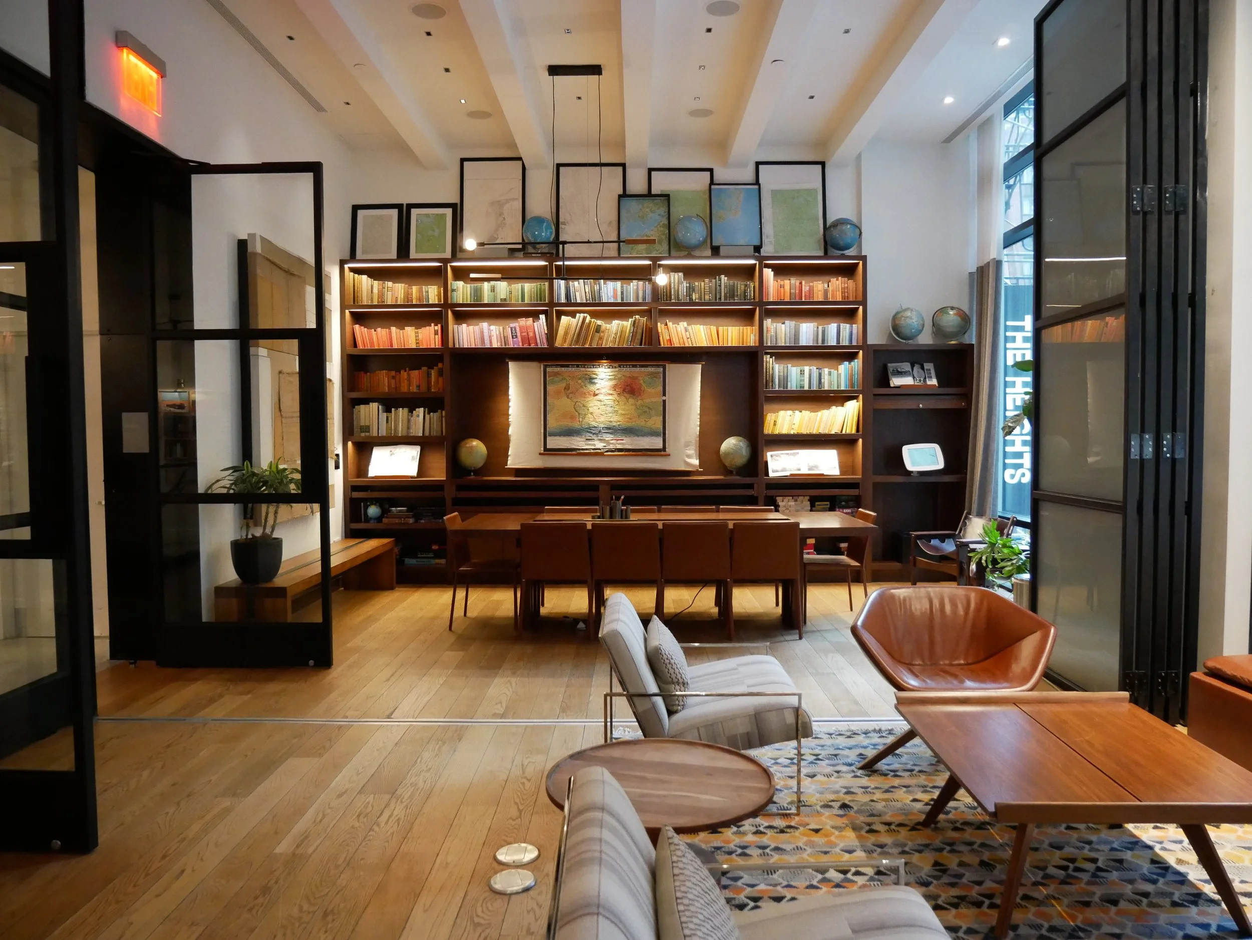 Une bibliothèque avec des étagères remplies de livres, un globe terrestre, un tableau d'art, et un coin salon avec fauteuils et tables en bois, éclairée par de grandes fenêtres.