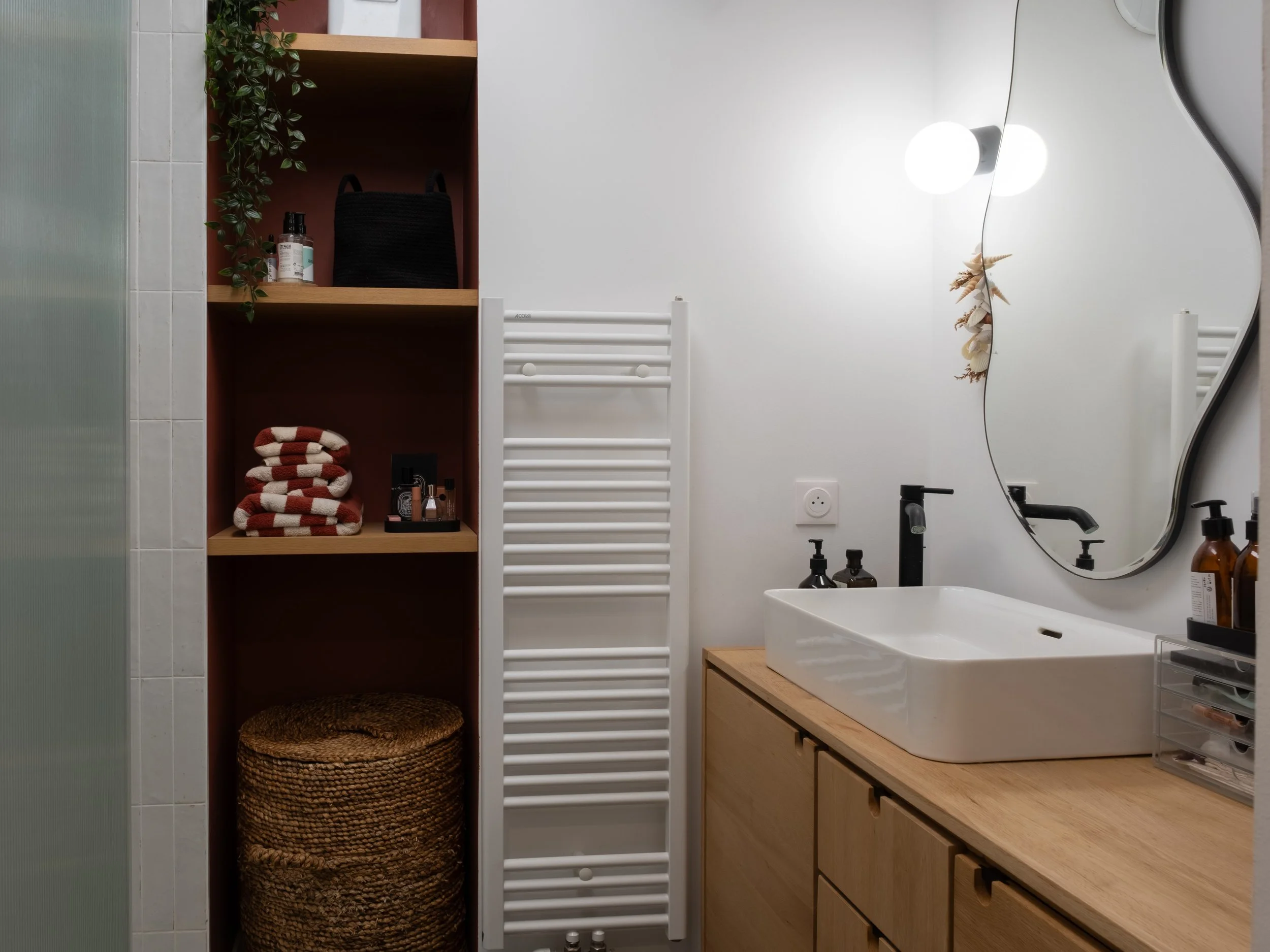 Un lavabo blanc sur un meuble en bois clair, avec un miroir incurvé au-dessus, dans une salle de bain moderne.