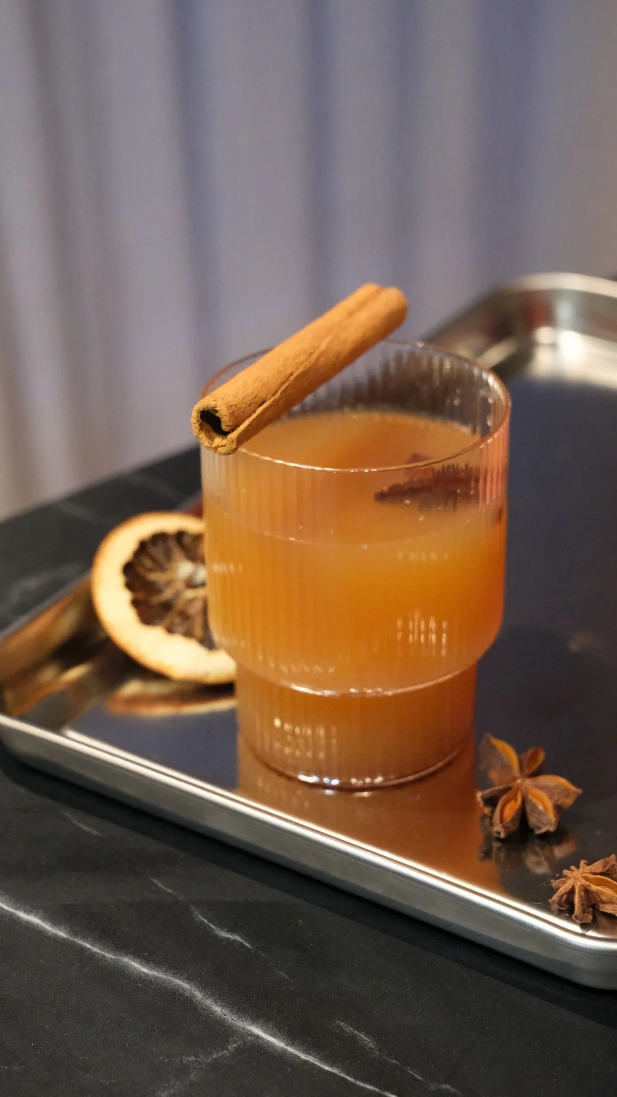 Verre à cocktail avec cannelle, une tranche d'orange séchée, clous de girofle, étoile de badiane et un plateau en métal noir.
