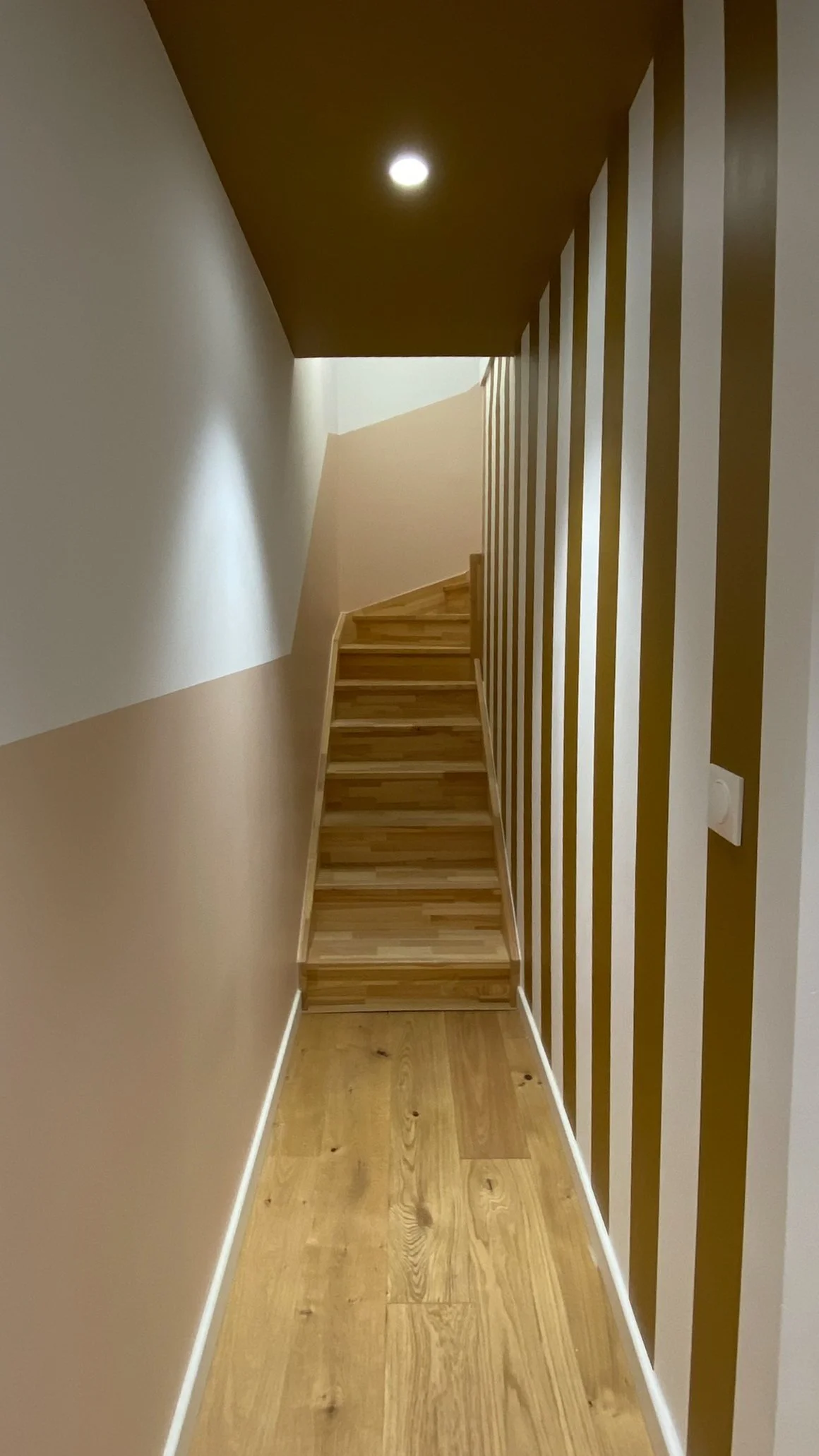 Une rampe d'escalier en bois dans une maison moderne avec un plafond en bois foncé et un mur avec des rayures verticales beige et blanches réalisées avec la peinture de marque Ressource teinte l'Alchimiste, un beau ocre lumineux.
