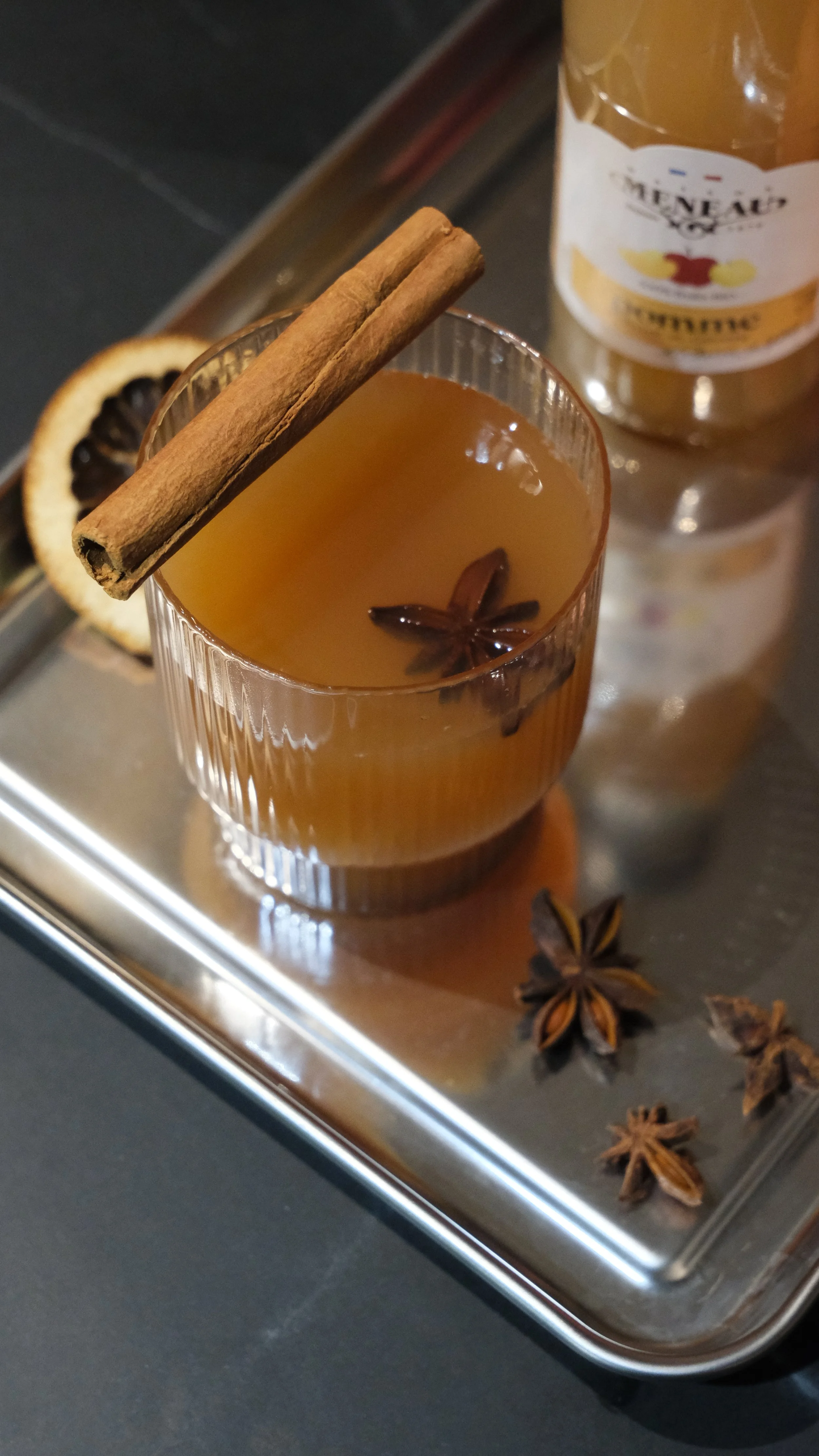 Verre de boisson chaude avec bâton de cannelle et étoile de badiane, décorée d’un rondelle de citron séchée sur un plateau en métal.