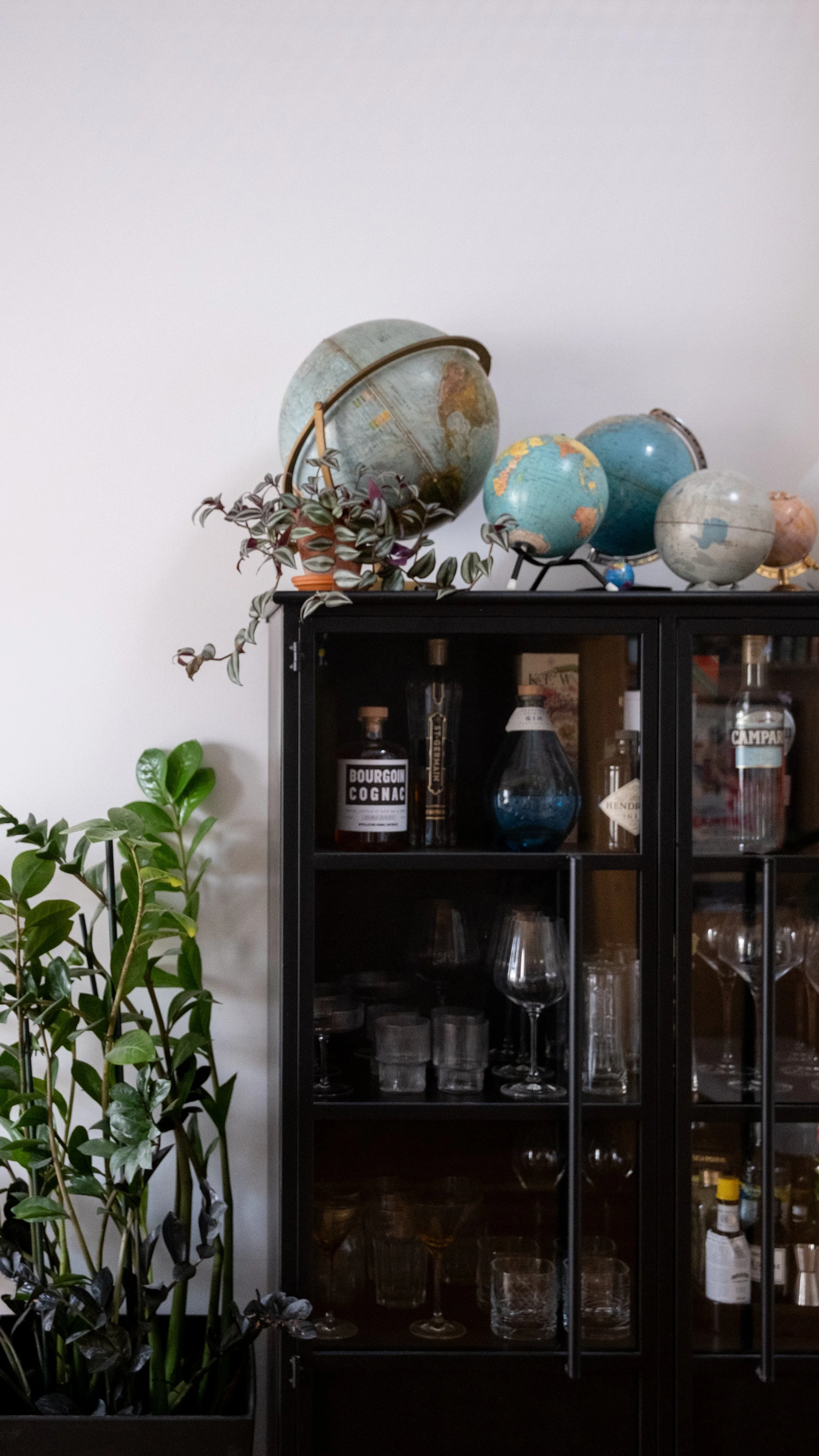 Une armoire noire en verre contenant des verres à vin et des bouteilles, surmontée de plusieurs globes terrestres vintage, un pot de plante verte, et un objet en forme de vase bleu.