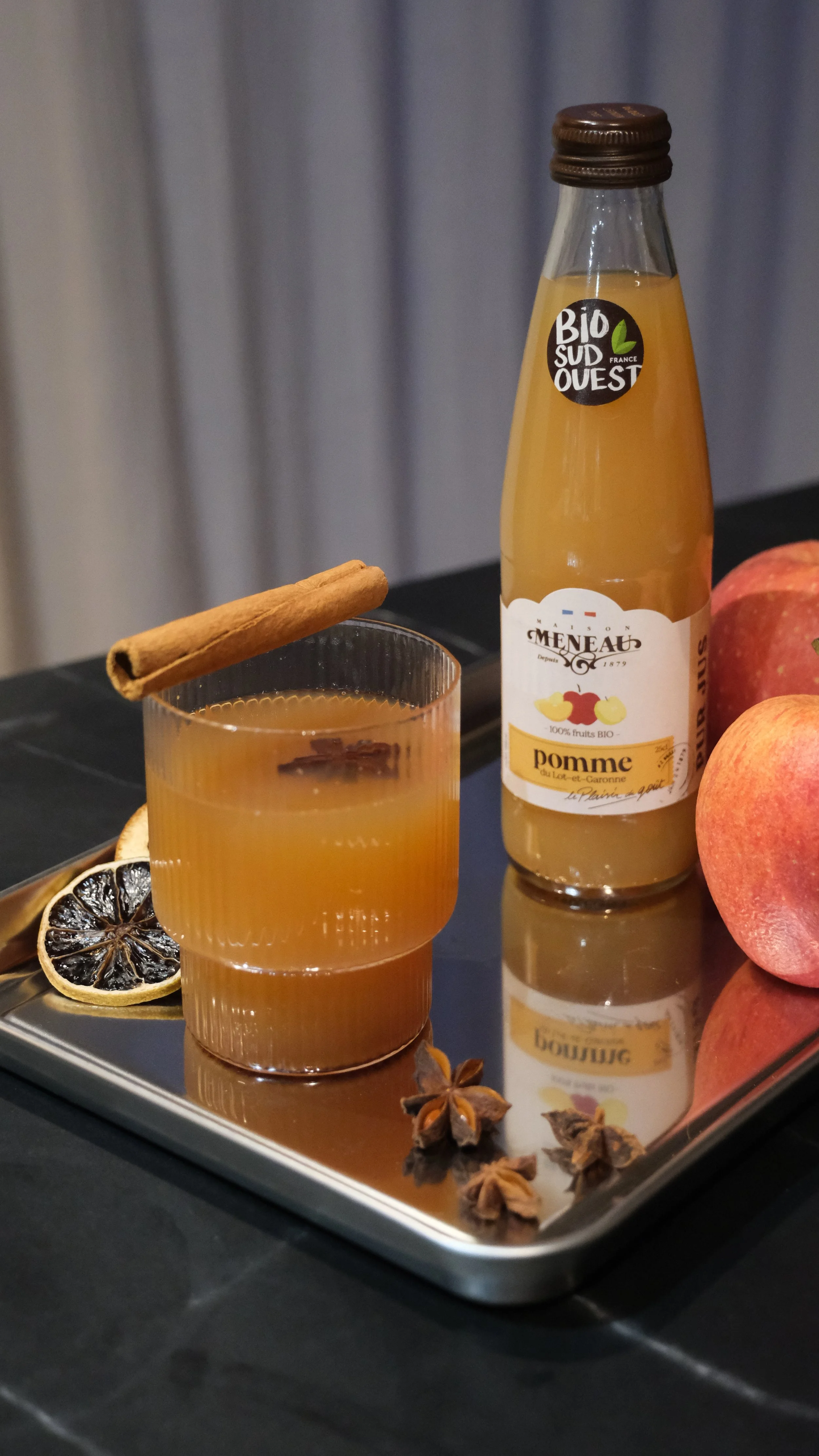 Verre de jus de pomme bio avec cannelle, étoile de badiane, tranches de citron noirci, sur une plaque en métal avec des pommes. Bouteille de jus de pomme et des pommes à côté.