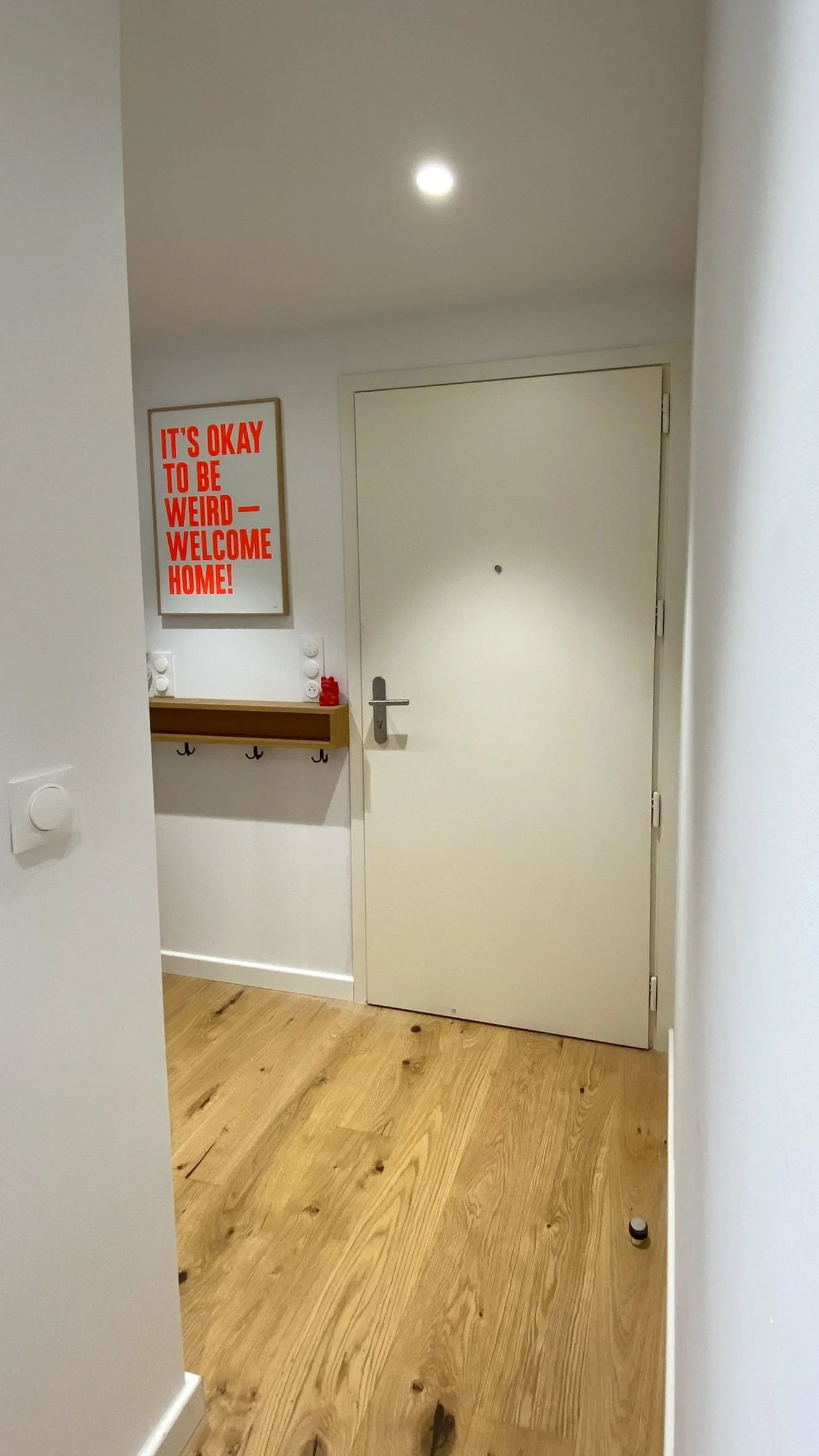 Entrée d'une pièce avec un mur blanc, un sol en bois chêne clair, une porte blanche, un tableau avec le texte en rouge "It's okay to be weird - Welcome home!", un petit étagère en bois avec des crochets. photo avant réalisation de la décoration
