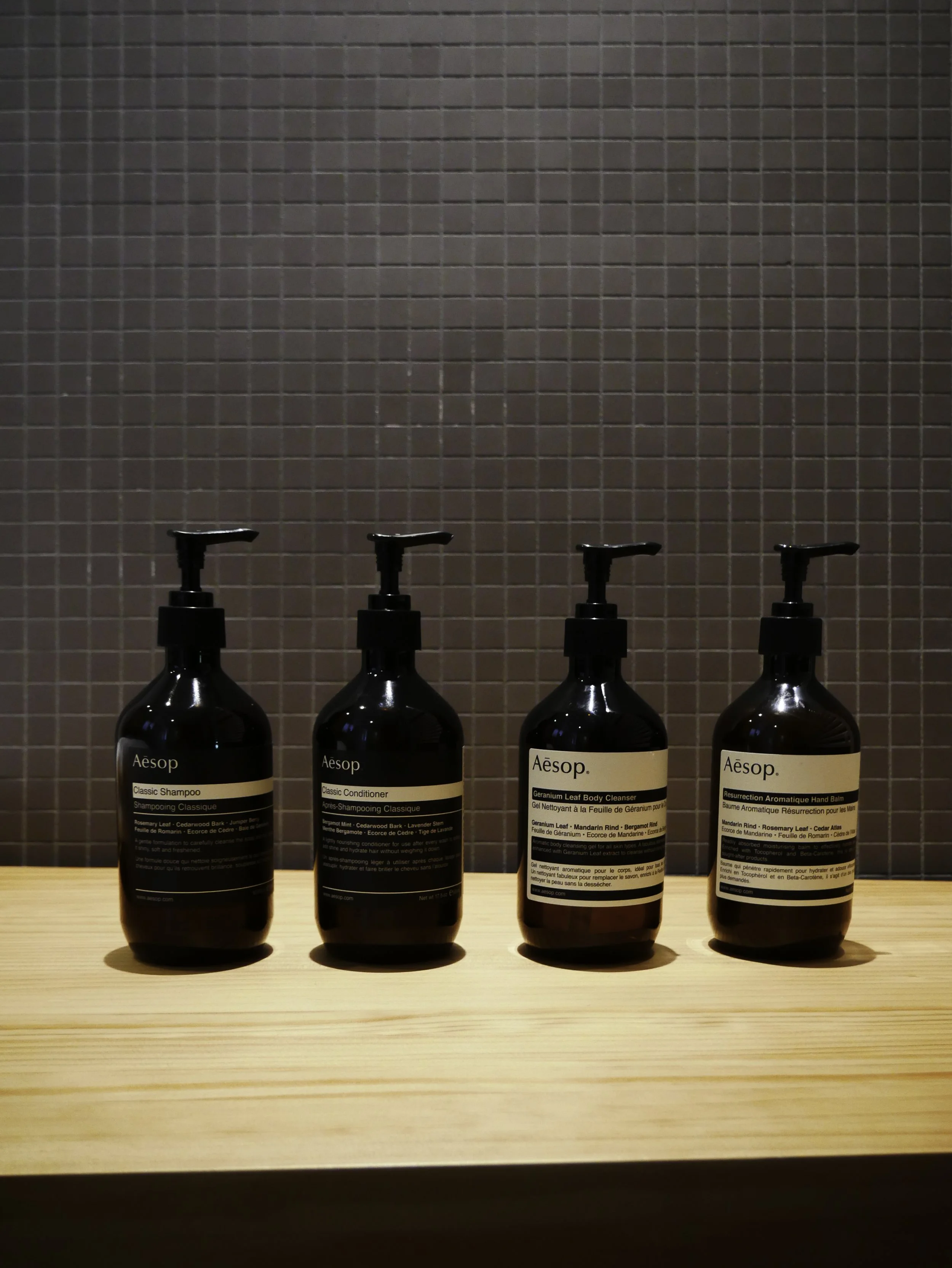 Quatre bouteilles de produits de soin Aesop avec pompe, alignées sur une étagère en bois contre un mur de carreaux gris.