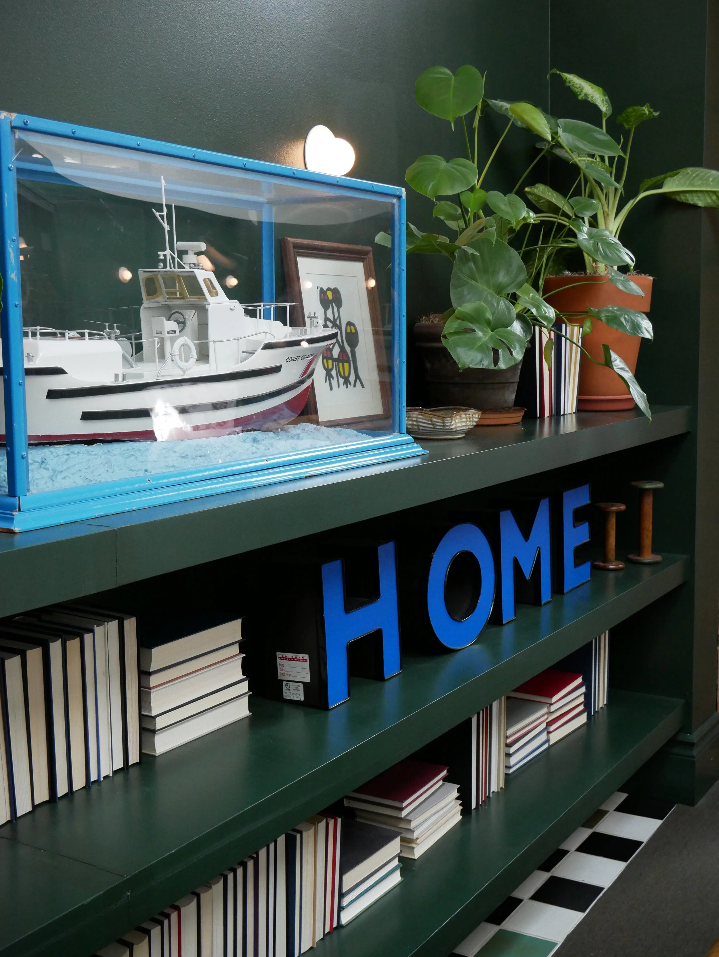 Une étagère verte avec des livres empilés, des plantes en pot et une pancarte bleue formant le mot "HOME". Sur le dessus, une maquette de bateau dans une vitrine et une photo encadrée d'un chien.