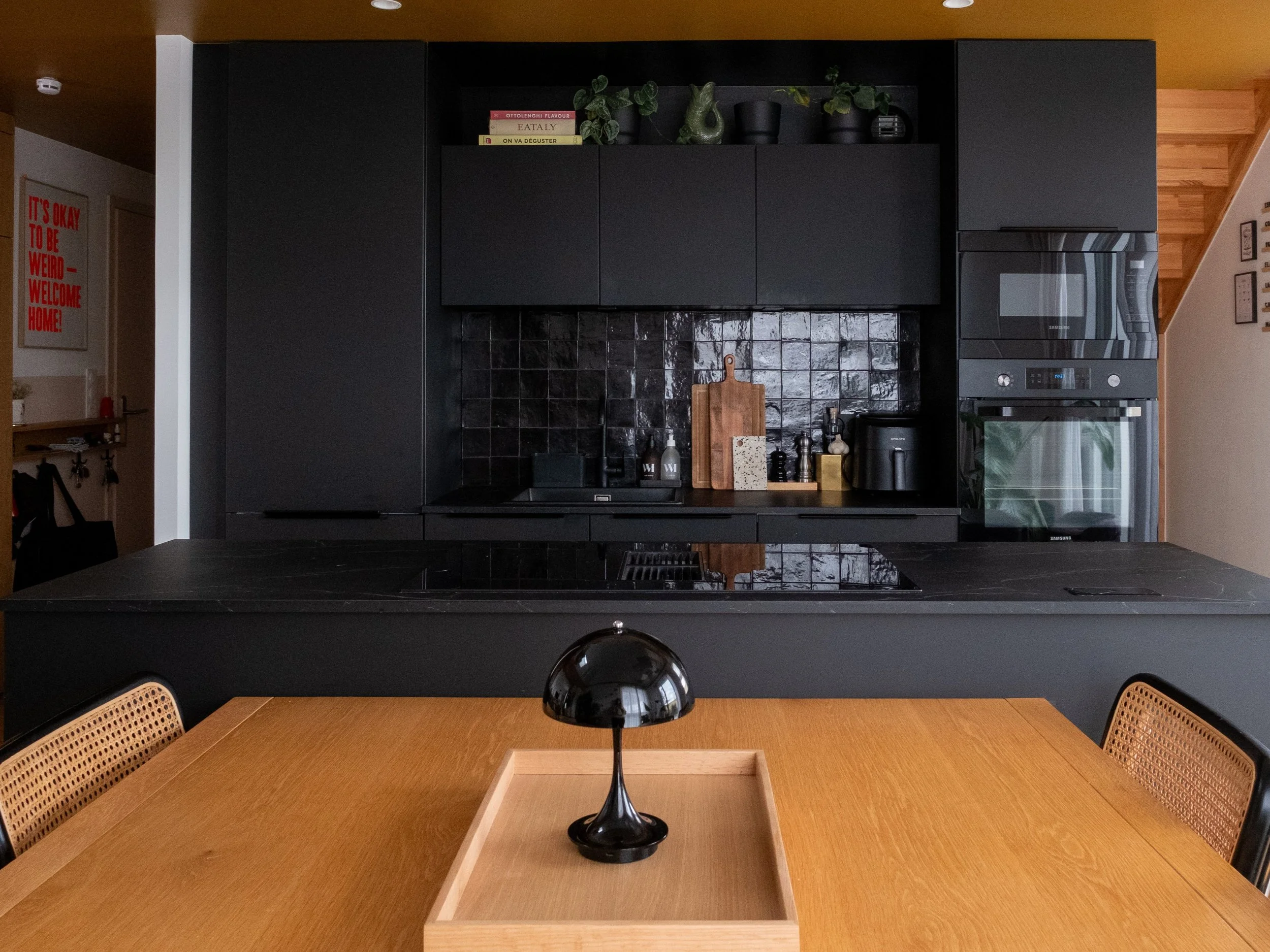 Cuisine moderne Schmidt avec une table en bois claire, lampe noire centrale sur un plateau en bois, et une cuisine noire avec des armoires, un dosseret en carreaux noirs, et un four encastré.