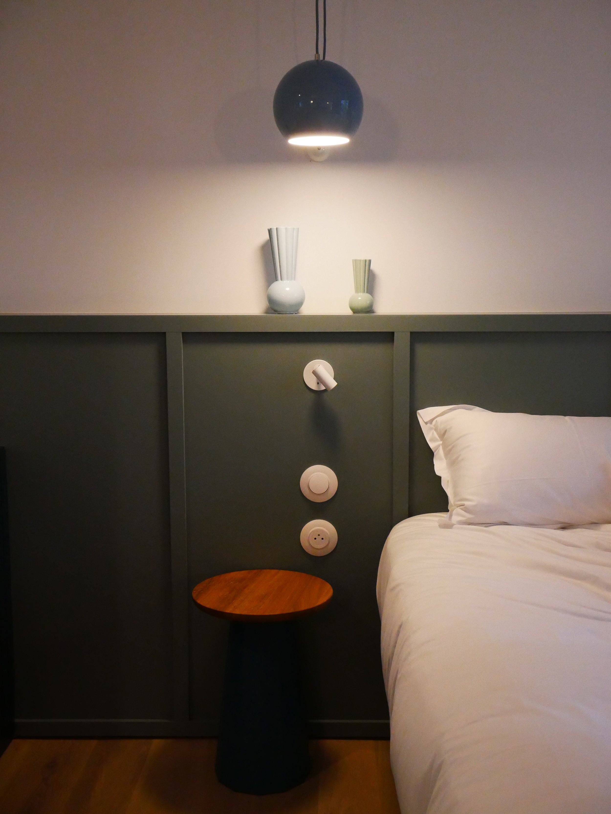 Lampadaire suspendu au plafond, deux vases avec des fleurs, un mur verdâtre avec des boutons et une lampe de lecture, un lit avec une taie d'oreiller blanche.