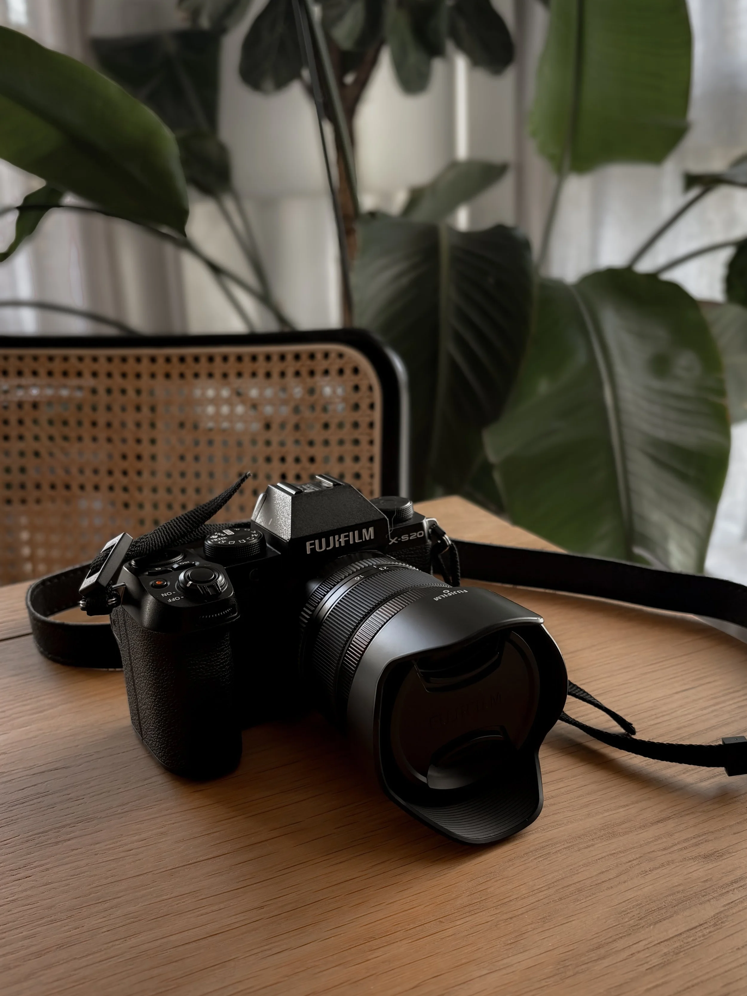 Appareil photo numérique Fujifilm X-S20 avec objectif sur une table en bois, arrière-plan avec des plantes vertes et une chaise en rotin.