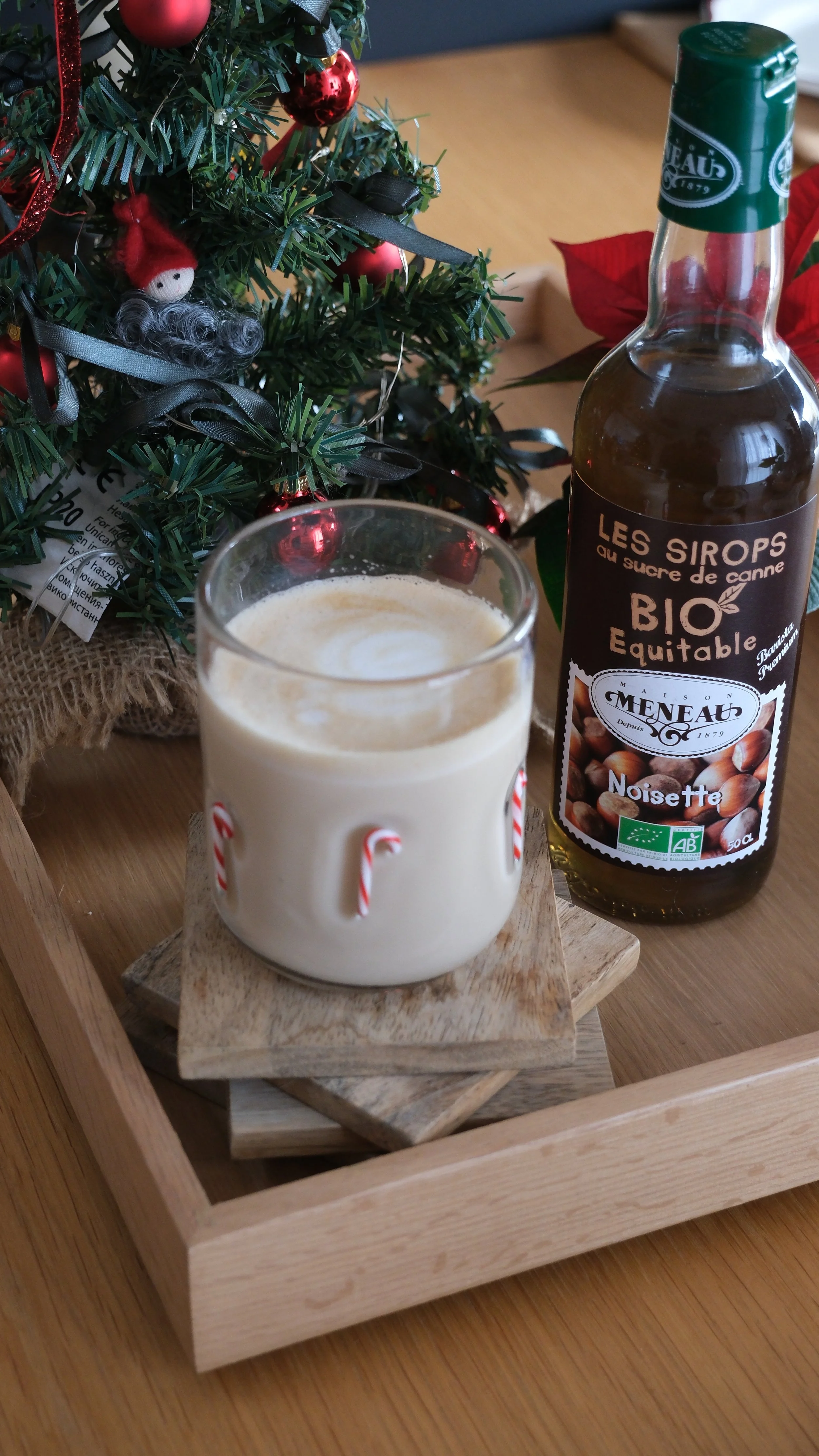 Une bouteille de sirop de noisette biologique à côté d'un verre de lait chaud orné de petits bonbons de Noël, avec un sapin de Noël décoré en arrière-plan.