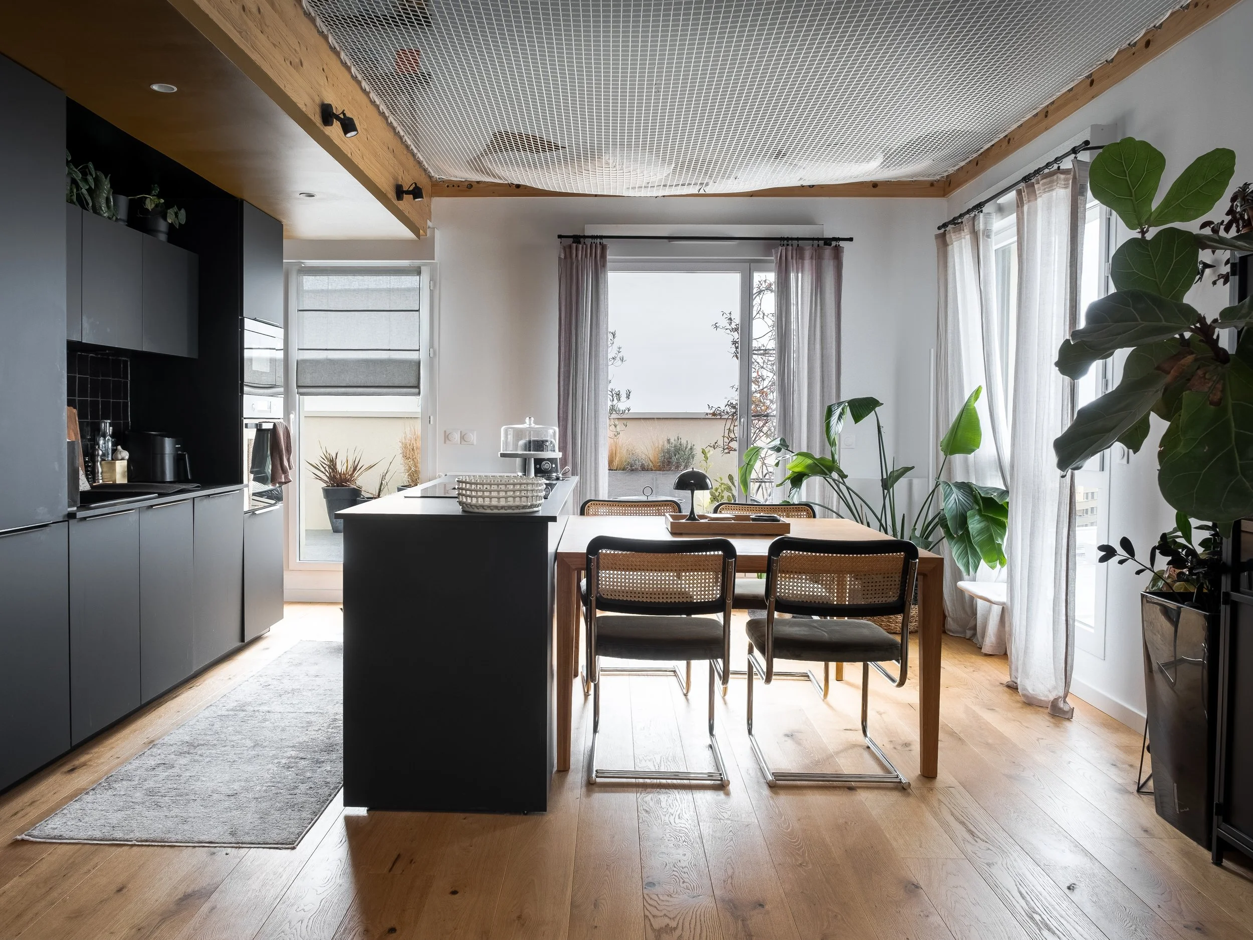 Intérieur d'une cuisine et salle à manger moderne avec des meubles noirs, une table en bois et des chaises, grandes fenêtres avec rideaux blancs, plusieurs plantes vertes, sol en bois, ambiance lumineuse naturelle.