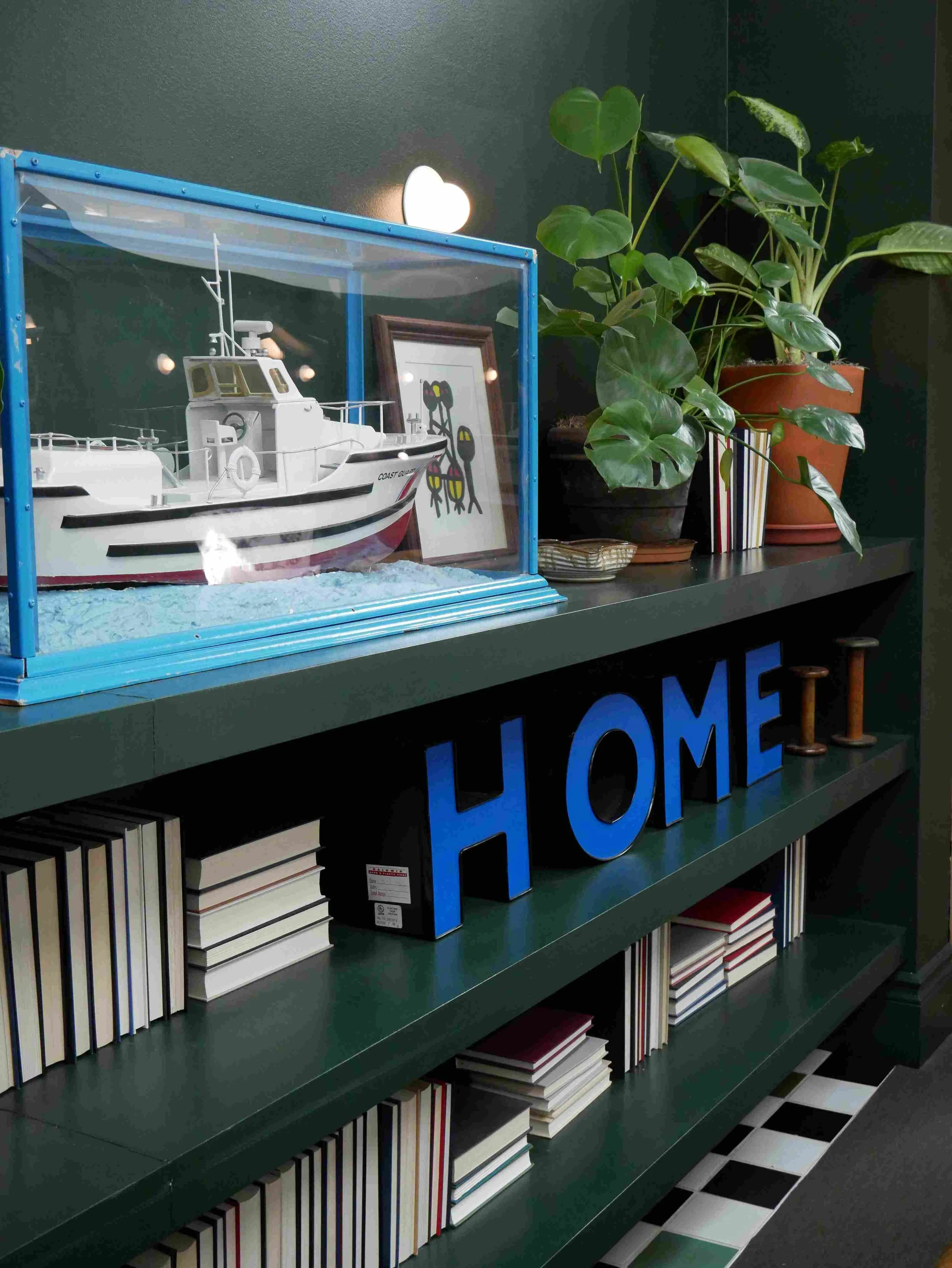 Une étagère verte avec des livres rangés verticalement, un grand mot en lettres bleues "HOME" sur l'étagère, et des plantes en pots derrière. Reportage photo professionnel d'hôtellerie.