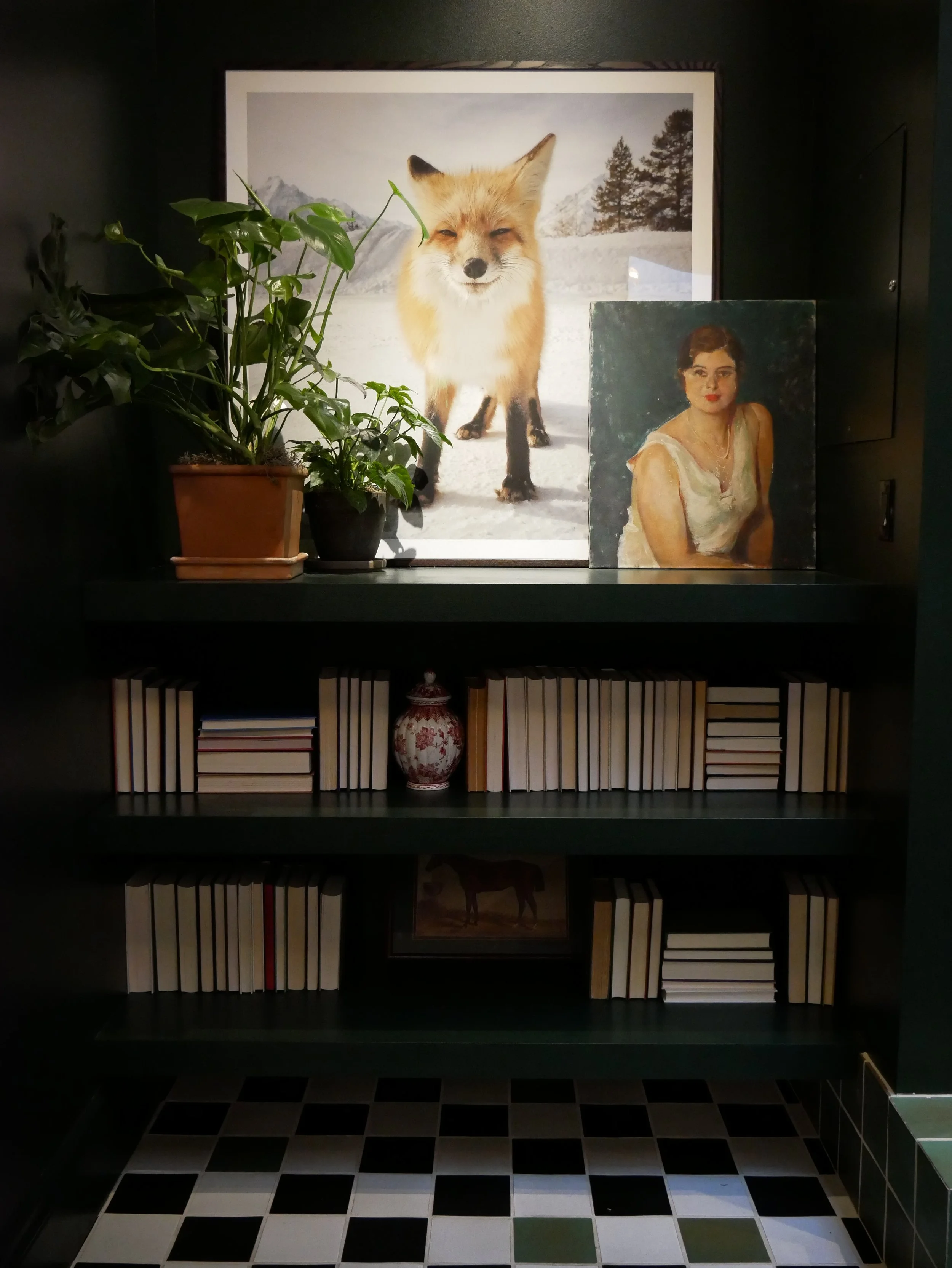 Une étagère sombre avec deux tableaux, un de renard dans la neige et un portrait de femme, entourée de livres empilés et une plante en pot à gauche.