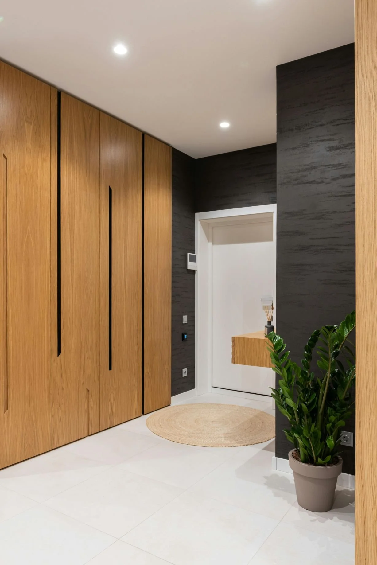 Intérieur moderne avec un mur en bois clair, un mur en pierre noire, un sol blanc, une porte blanche, un tapis rond en osier, une plante verte dans un pot gris et un petit meuble en bois. Proposition de décoration de parties communes immeuble copro