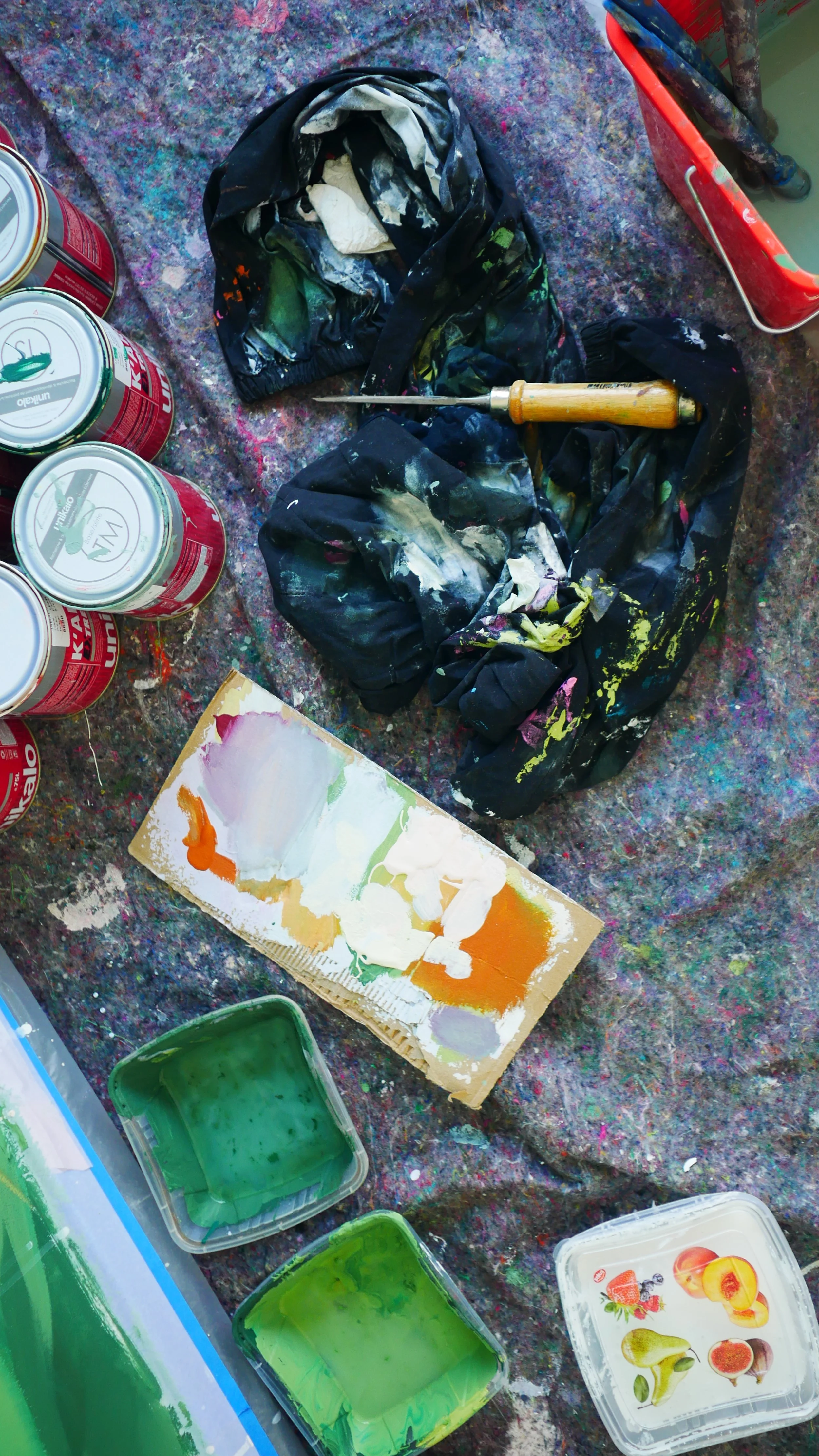 Espace de travail artistique avec peintures, pinceaux, contenants de peinture, et un tapis ou tissu imprimé, évoquant une scène de peinture ou de création artistique.