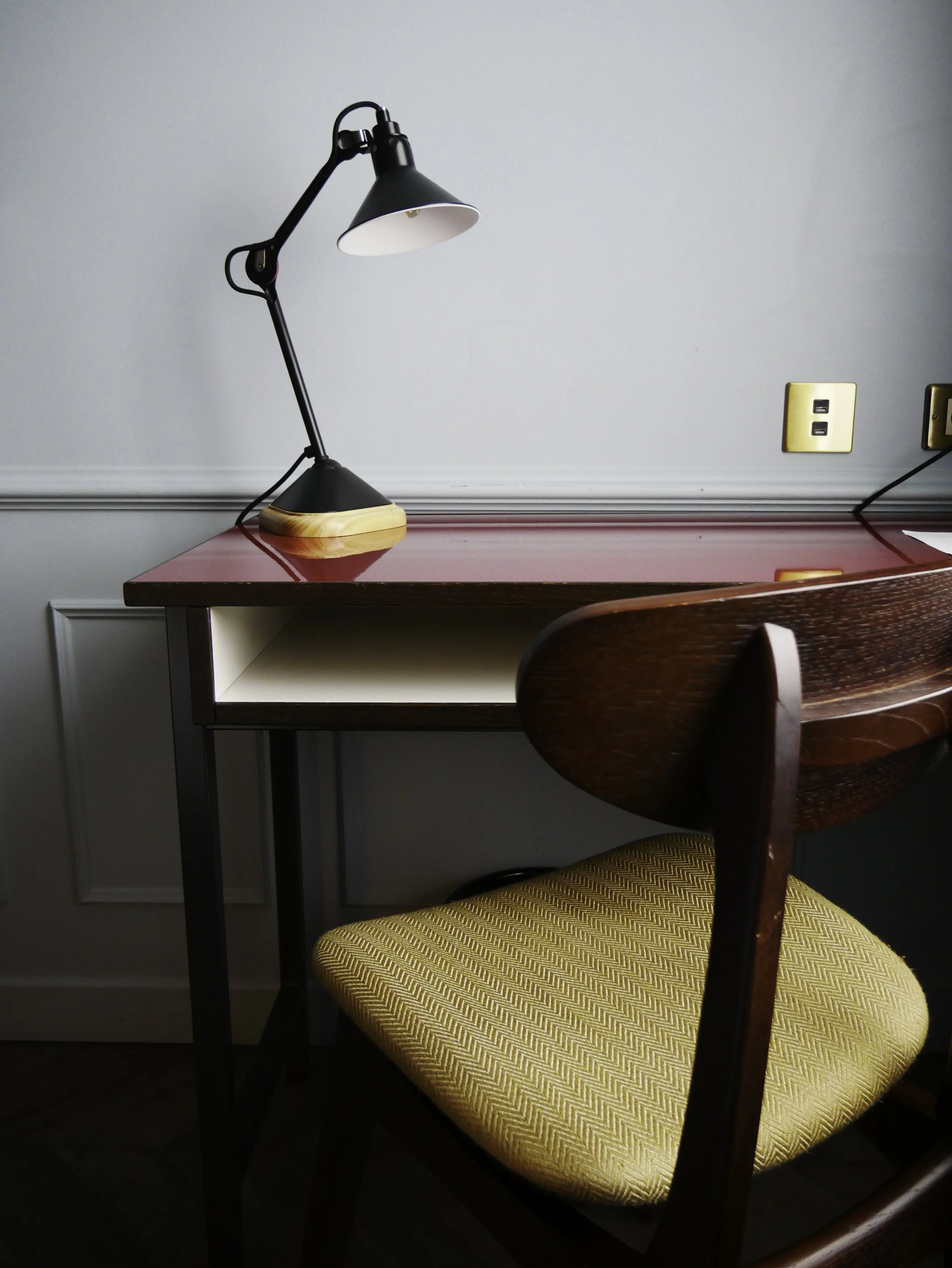 Un bureau avec une lampe noire, une prise électrique et un fauteuil en bois avec un coussin jaune. La surface du bureau est rouge.