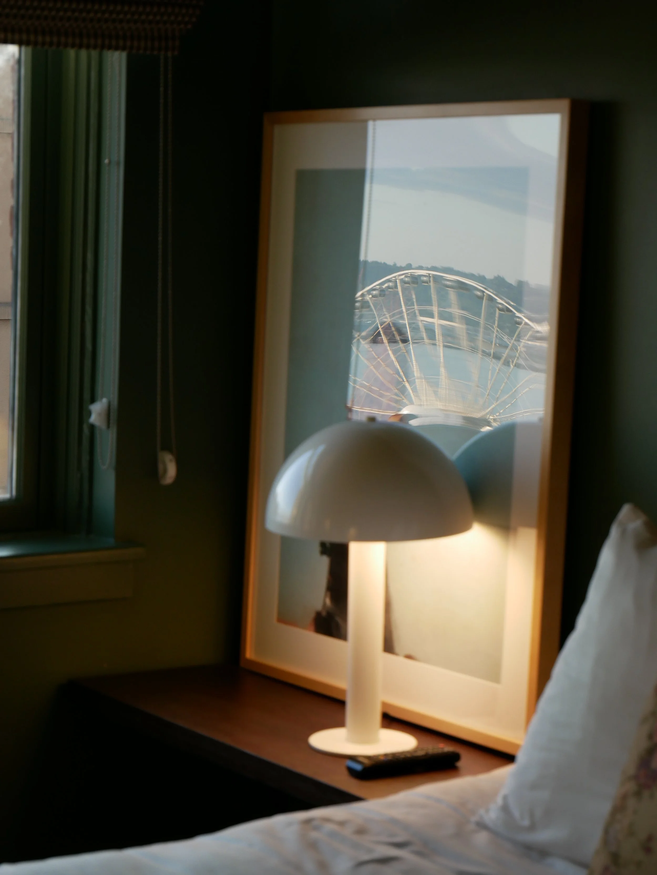 Une lampe de bureau blanche sur une table en bois, avec un miroir derrière et une fenêtre à gauche, dans une chambre. On voit un lit à droite.
