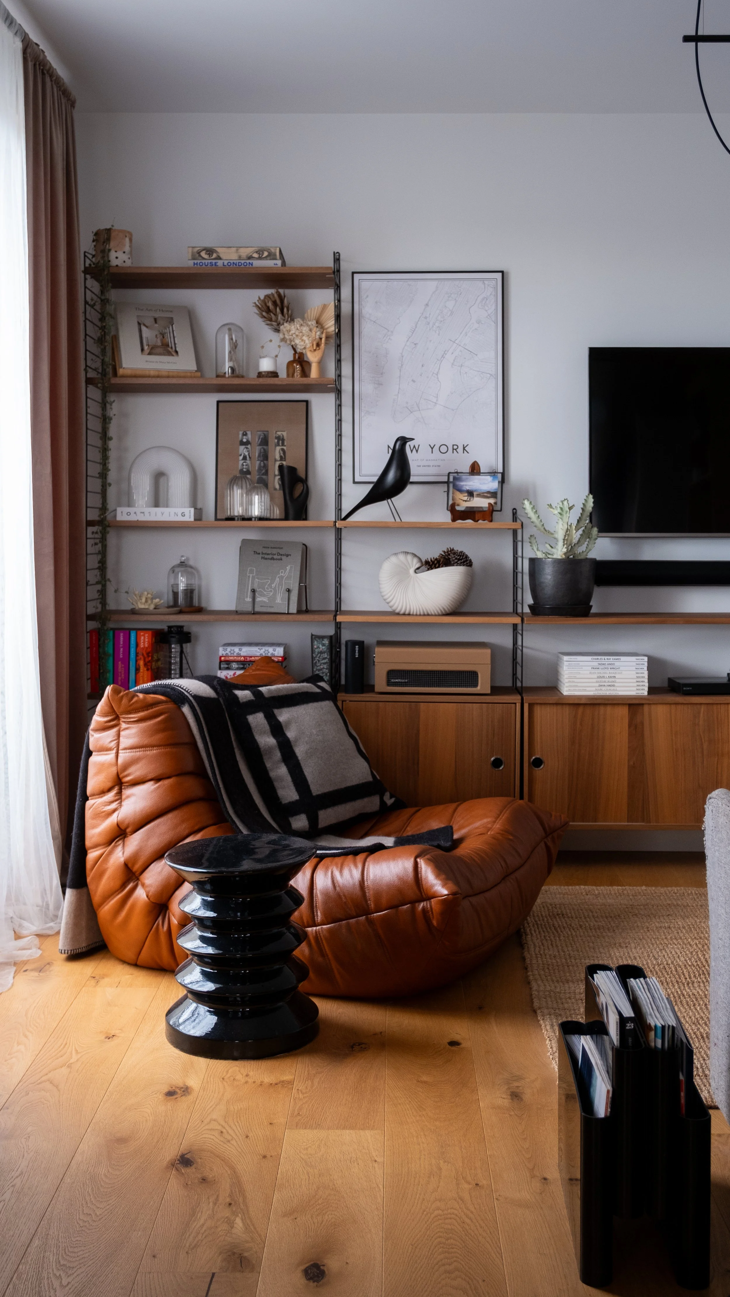 Salon moderne avec fauteuil en cuir marron Togo de Ligne Roset, étagère en bois et métal String Furniture, décorations telles que des livres, des plantes, des sculptures et un tableau de New York, avec un téléviseur mural