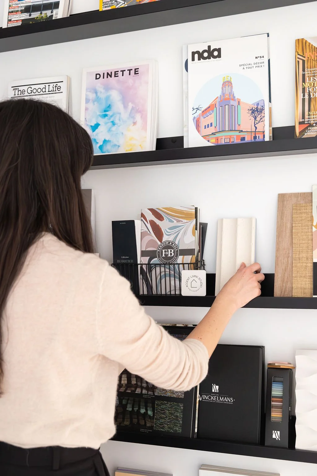 Une décoratrice d'intérieur à Bordeaux, portant une chemise beige, regarde une étagère remplie de magazines de décoration, catalogues et échantillons de matériaux dans un intérieur showroom.