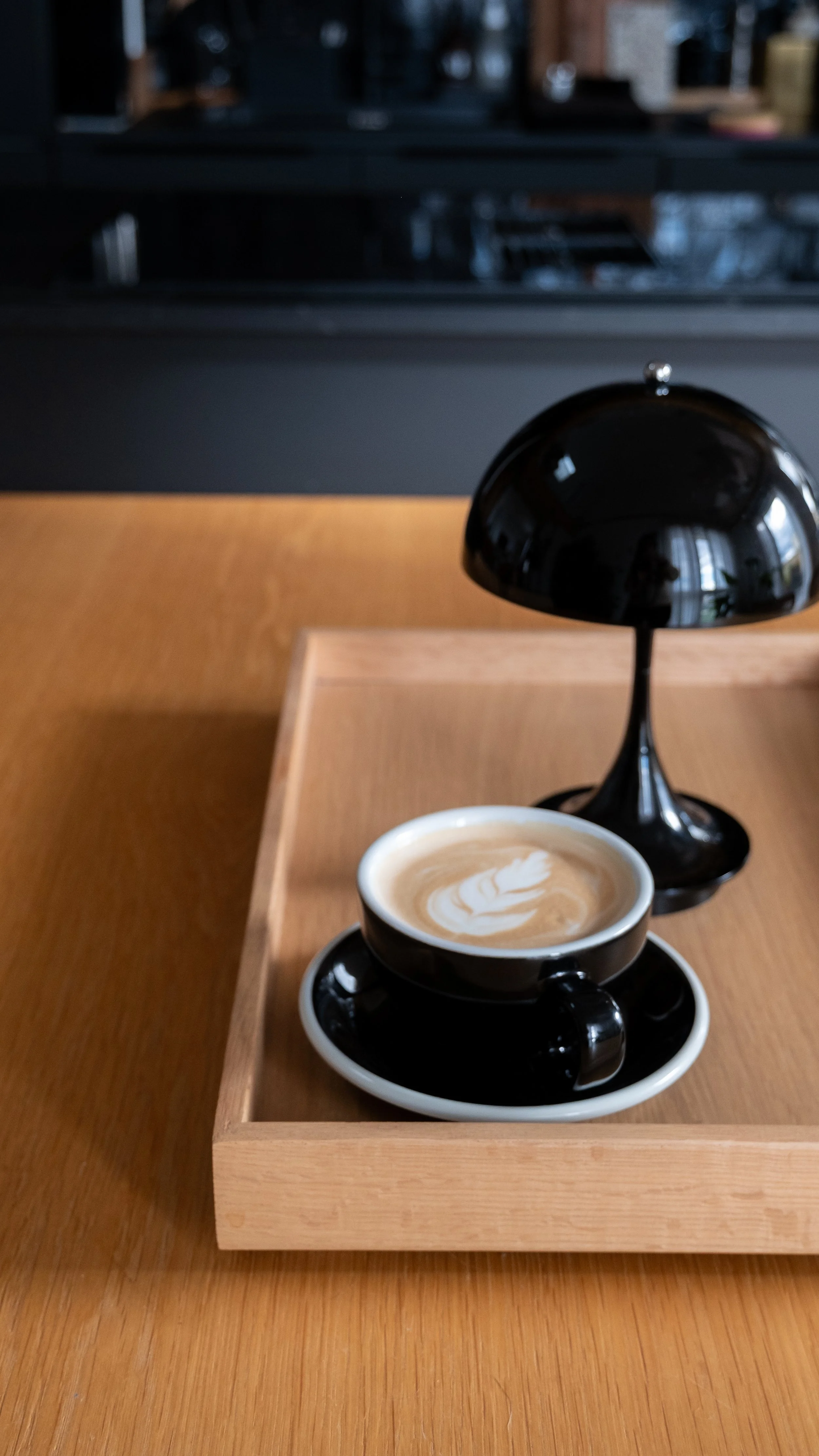 Une tasse Loveramics de café au lait avec un motif latte art sur le dessus, placée sur une soucoupe noire, avec une lampe Panthella de Louis Poulsen noir à côté, tous deux sur un plateau en bois sur une table en bois.