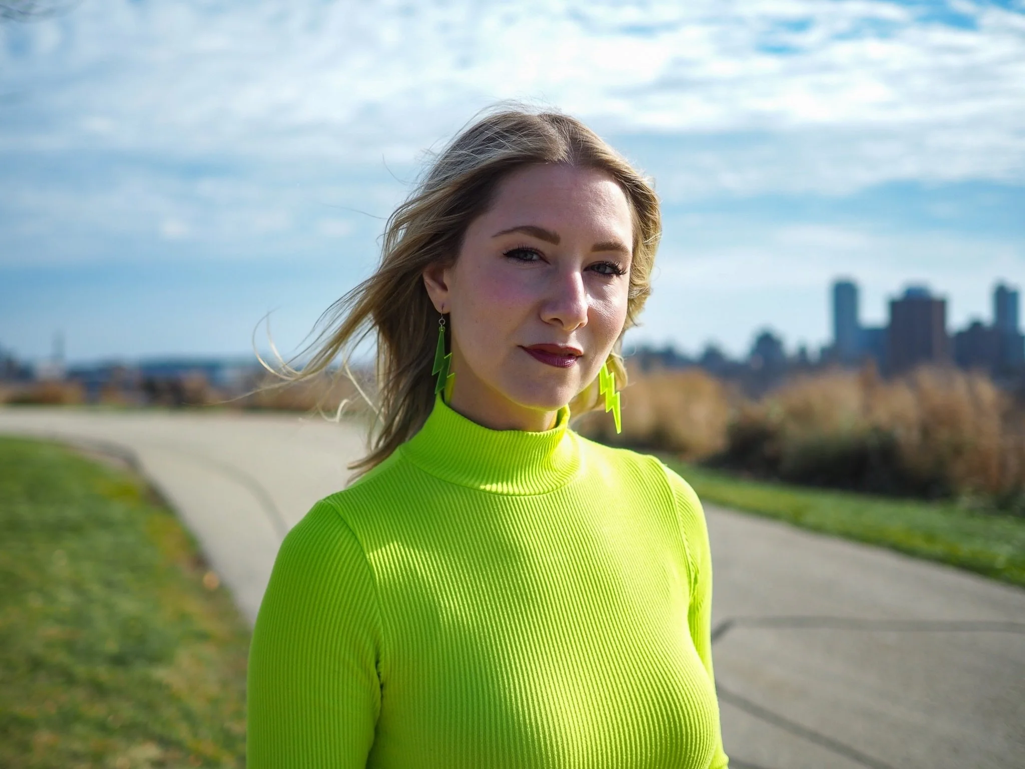 A woman with blonde hair wearing a bright yellow-green top and lightning bolt earrings standing outdoors on a walking trail with city buildings in the background.