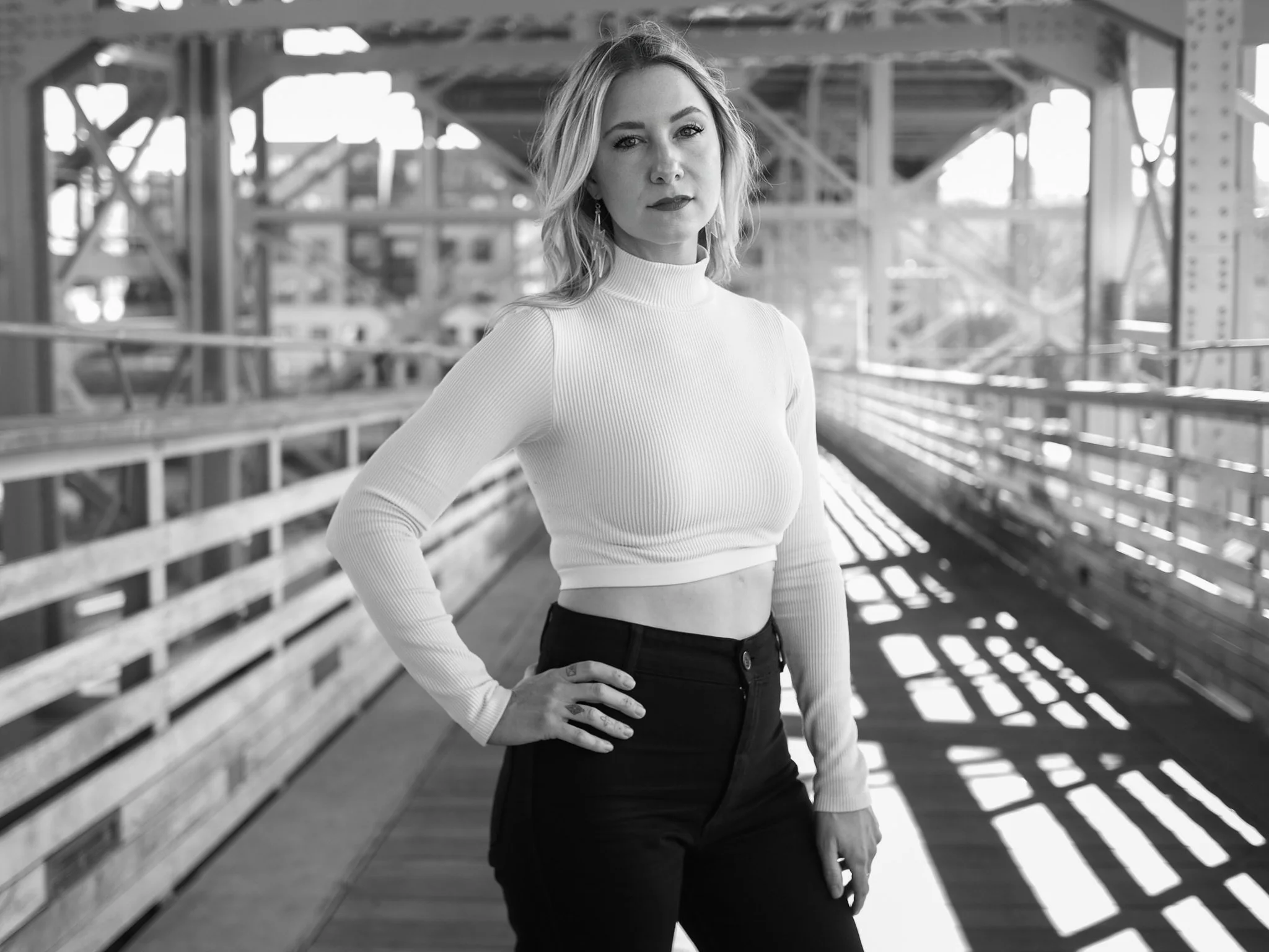 A woman with shoulder-length hair standing on a bridge or walkway with a metal structure, wearing a long-sleeve, high-neck crop top and high-waisted pants, looking at the camera with her hand on her hip.
