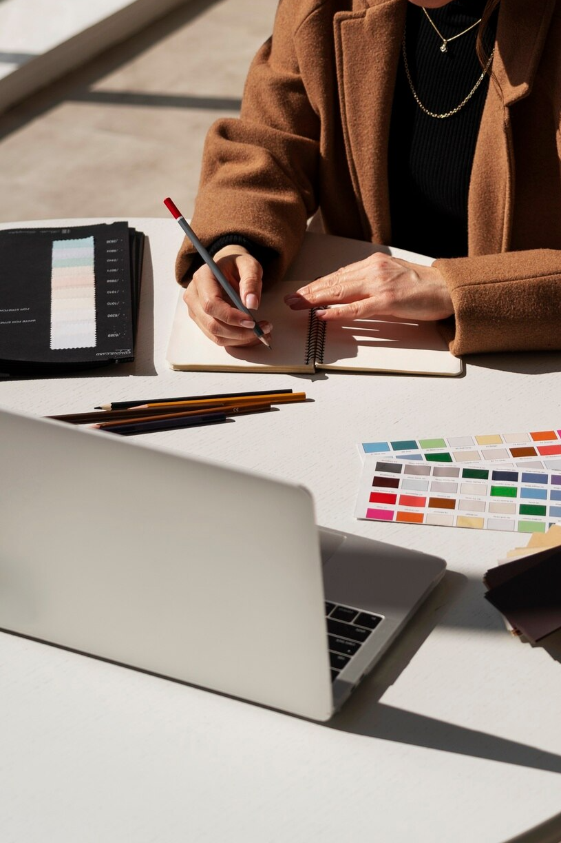 Person using a pen to make notes in a notebook at a workspace with color swatches, pencils, and a laptop.