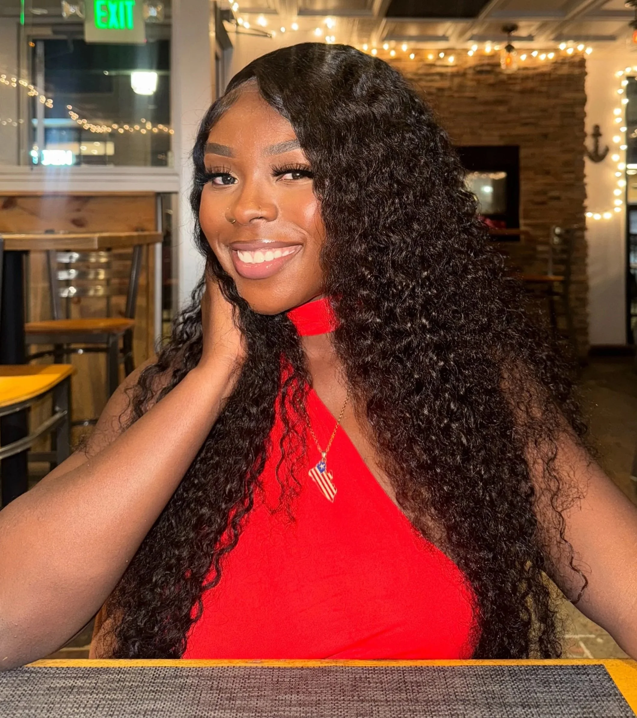 A woman smiling at the camera, wearing a red dress, in a restaurant with string lights and wooden decor.