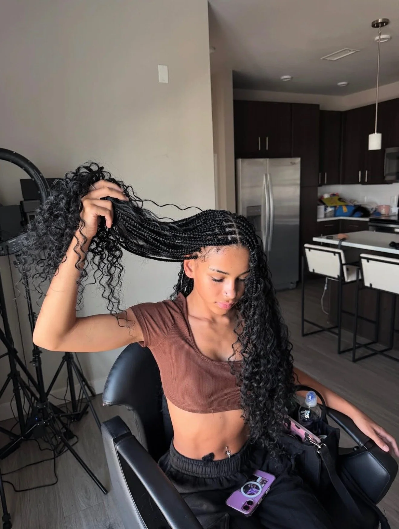 A woman with long curly hair sitting in a chair, holding a section of her hair with her hand, in a kitchen with dark cabinets and a refrigerator in the background.