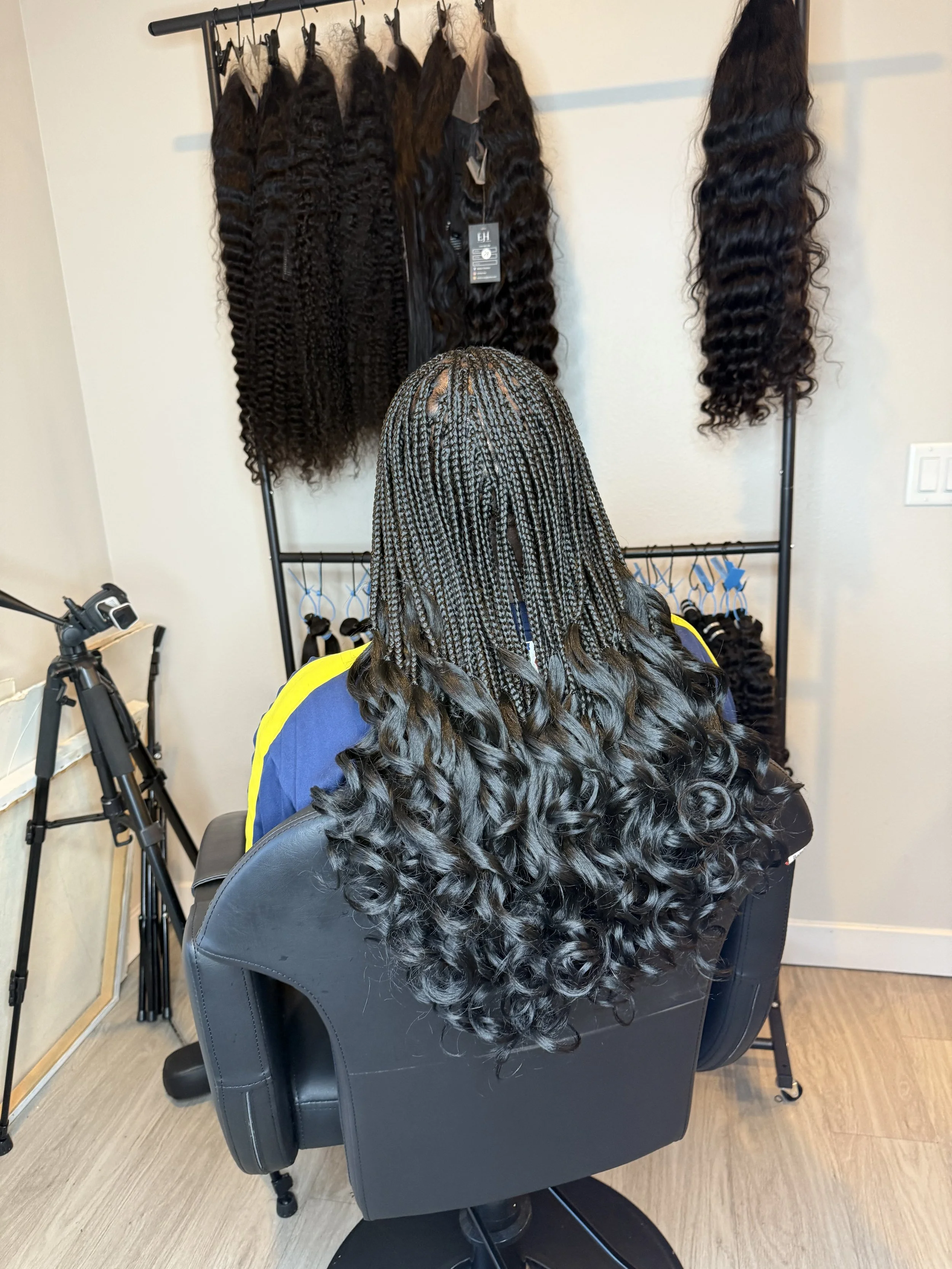 Rear view of a person with long black hair styled in tight curls, sitting in a salon chair, with hair extensions hanging on a rack behind them.