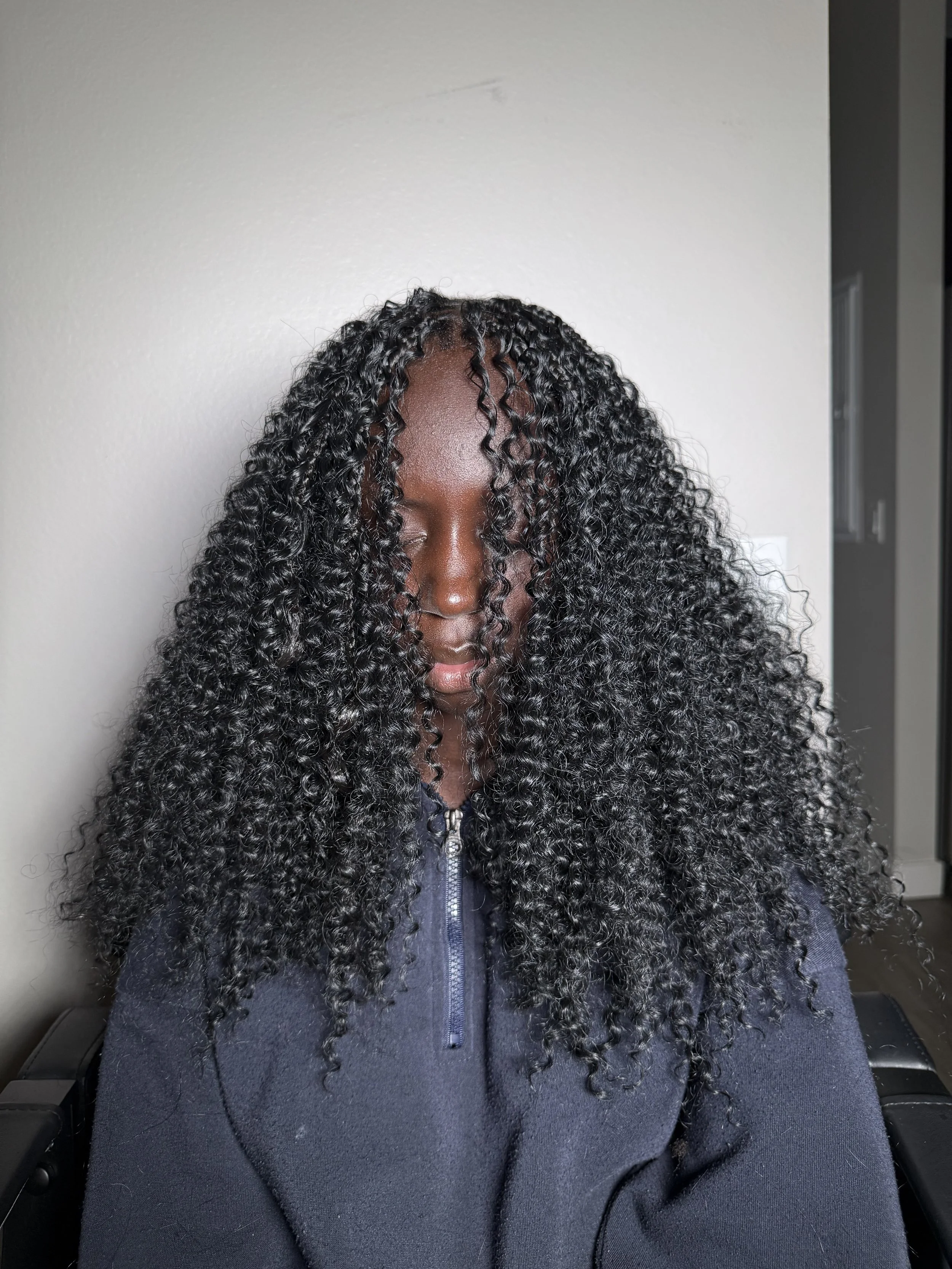 A woman with long, curly black hair covering her face, wearing a navy blue zip-up jacket, seated indoors against a plain wall.