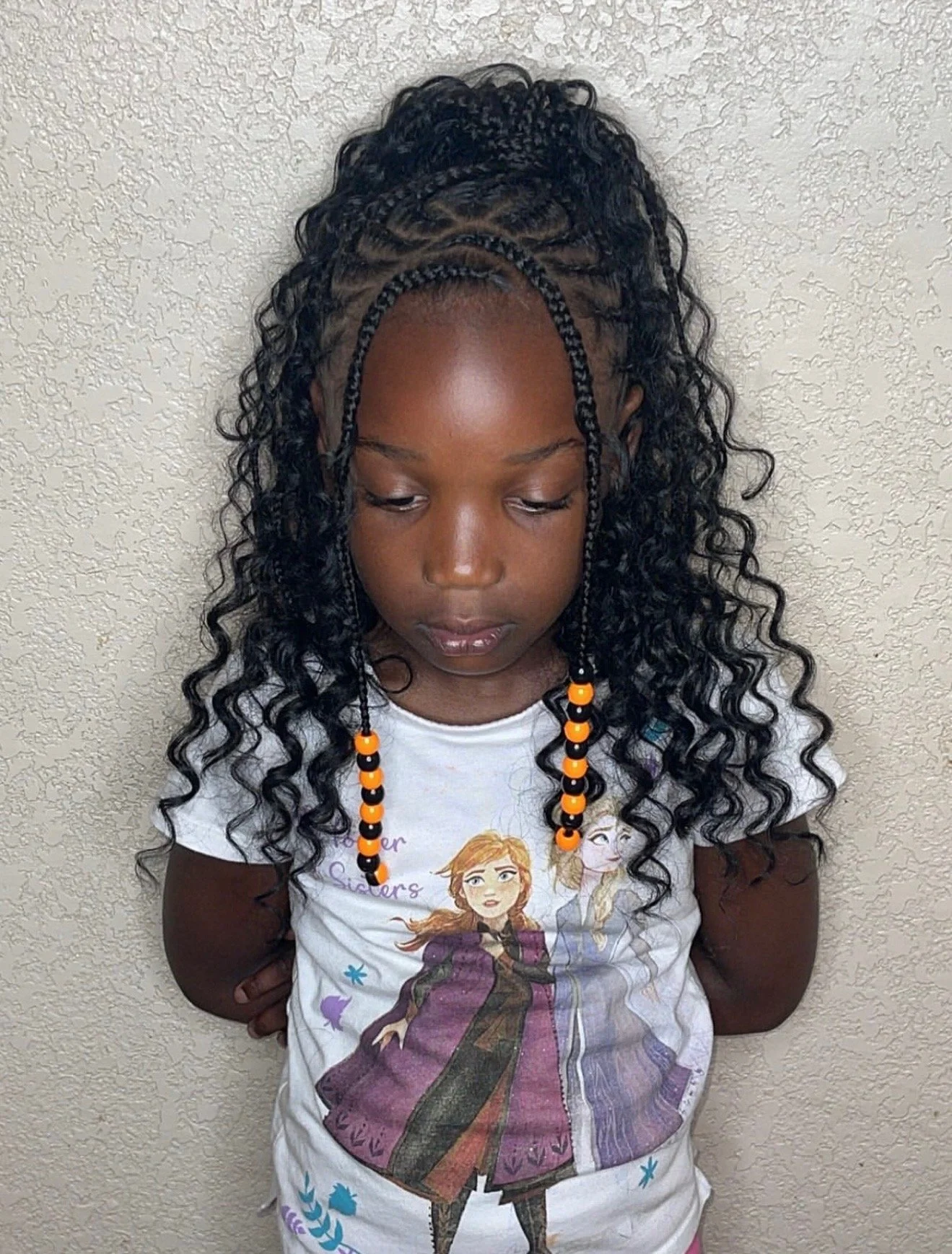 Young girl with dark, curly hair styled in braids with orange and black beads, wearing a "Frozen" t-shirt, standing against a textured wall with her head slightly tilted downward.