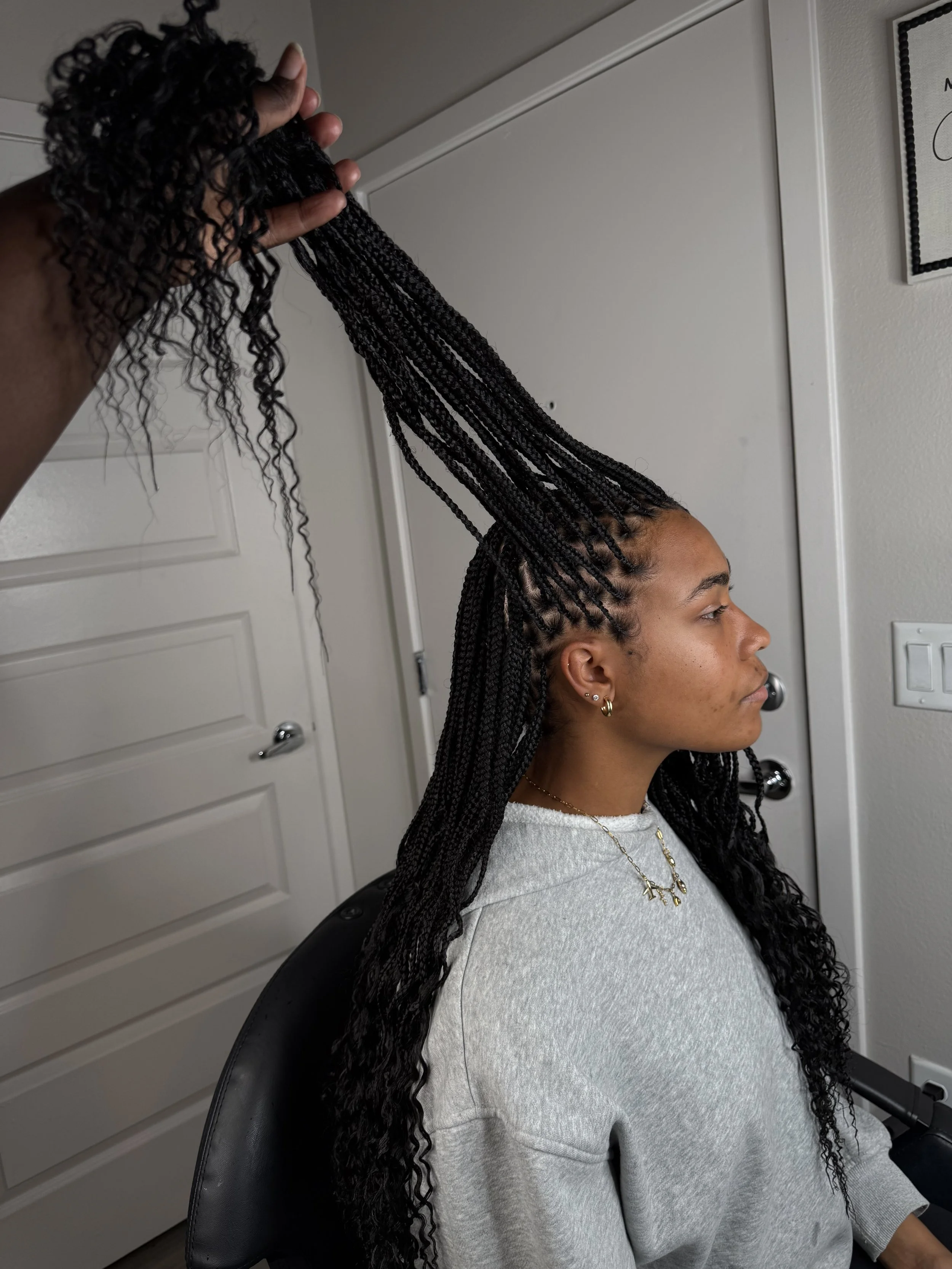 A person with long, dark, curly hair is having their hair styled into thin braids by a stylist. The person is seated in a salon chair, wearing a light gray sweatshirt and gold jewelry, with a neutral expression.
