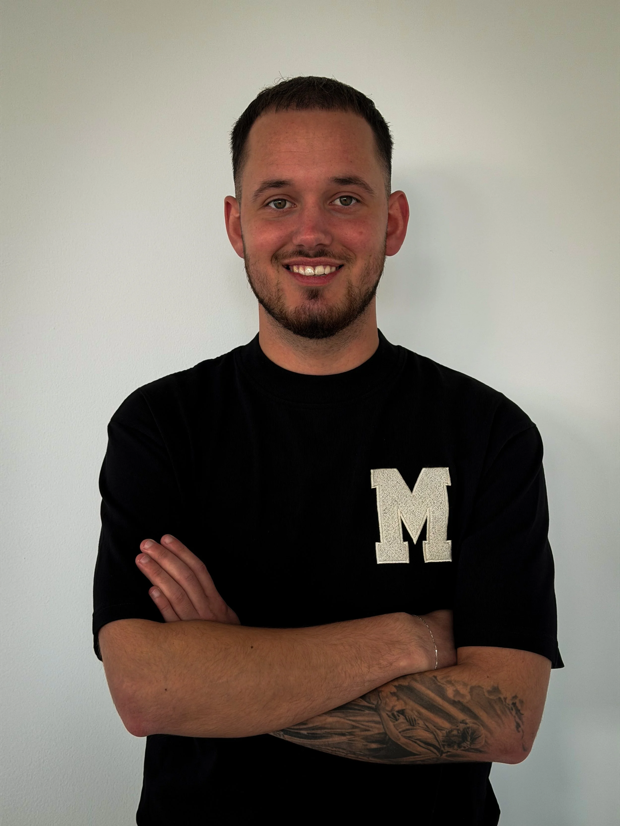 A young woman with shoulder-length black hair smiling outdoors, wearing a blue top, surrounded by green plants in a greenhouse or garden.