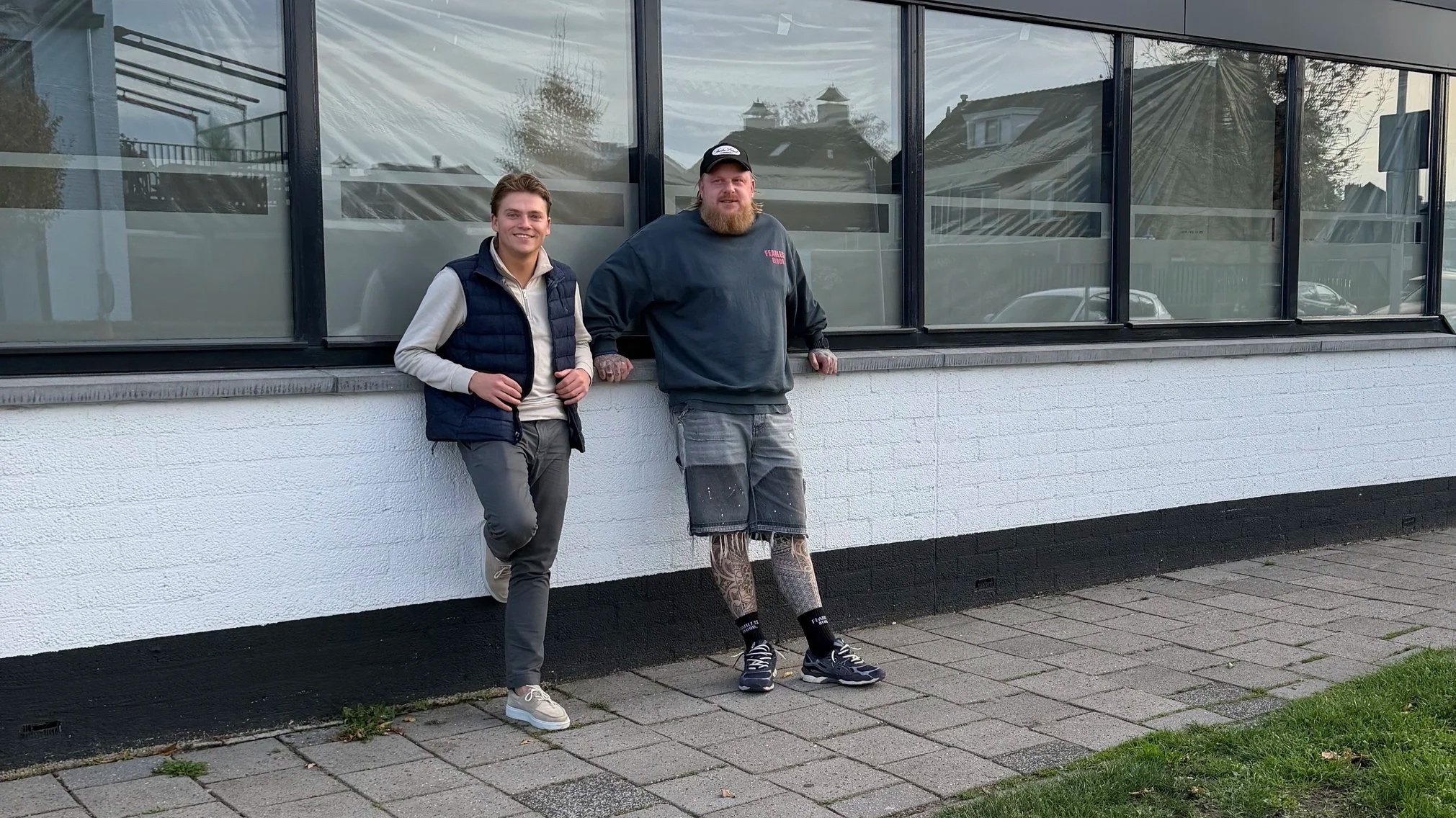 Two men stand outside a building with large window panels. One man is young, dressed casually in a vest, shirt, and pants. The other man is older, with a beard, wearing a sweatshirt, shorts, and has tattooed legs. They are smiling and leaning against the wall.