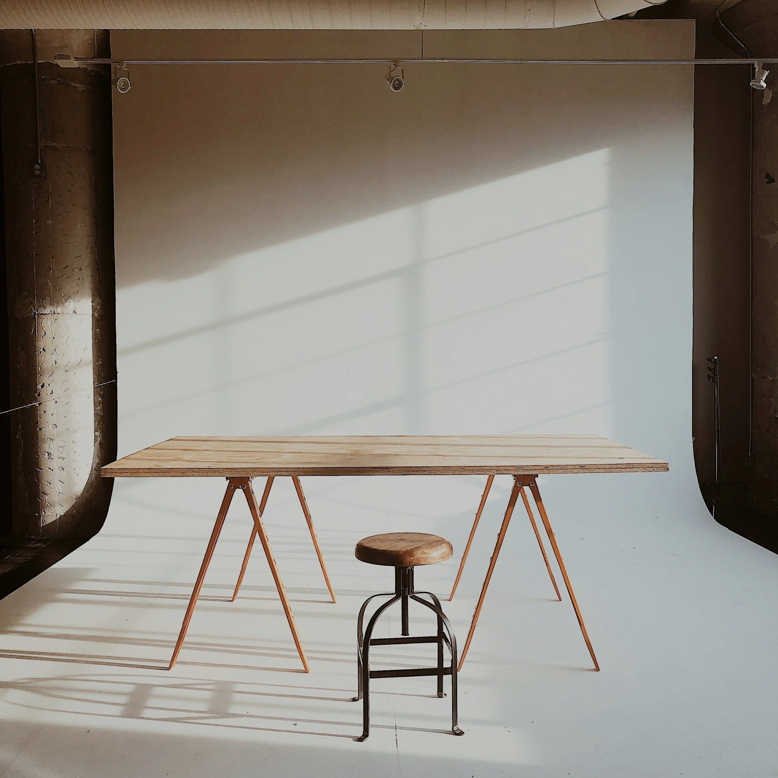 A wooden table with three A-frame legs, a small rustic stool with metal legs, and a bright backdrop with sunlight and shadows.
