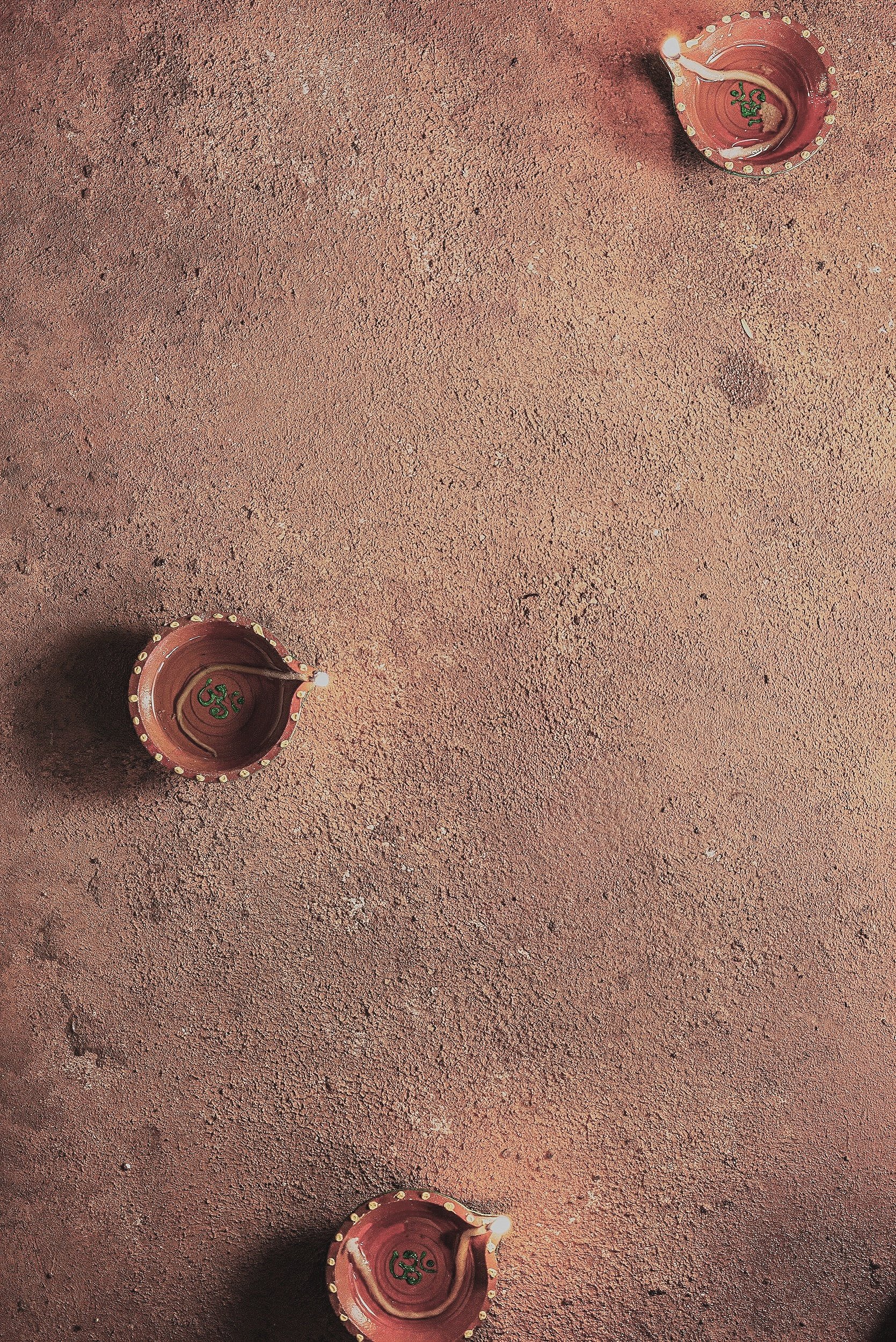 Three unlit clay diyas placed on a textured, reddish-brown surface, arranged in a scattered manner, with two at the top and one at the bottom.