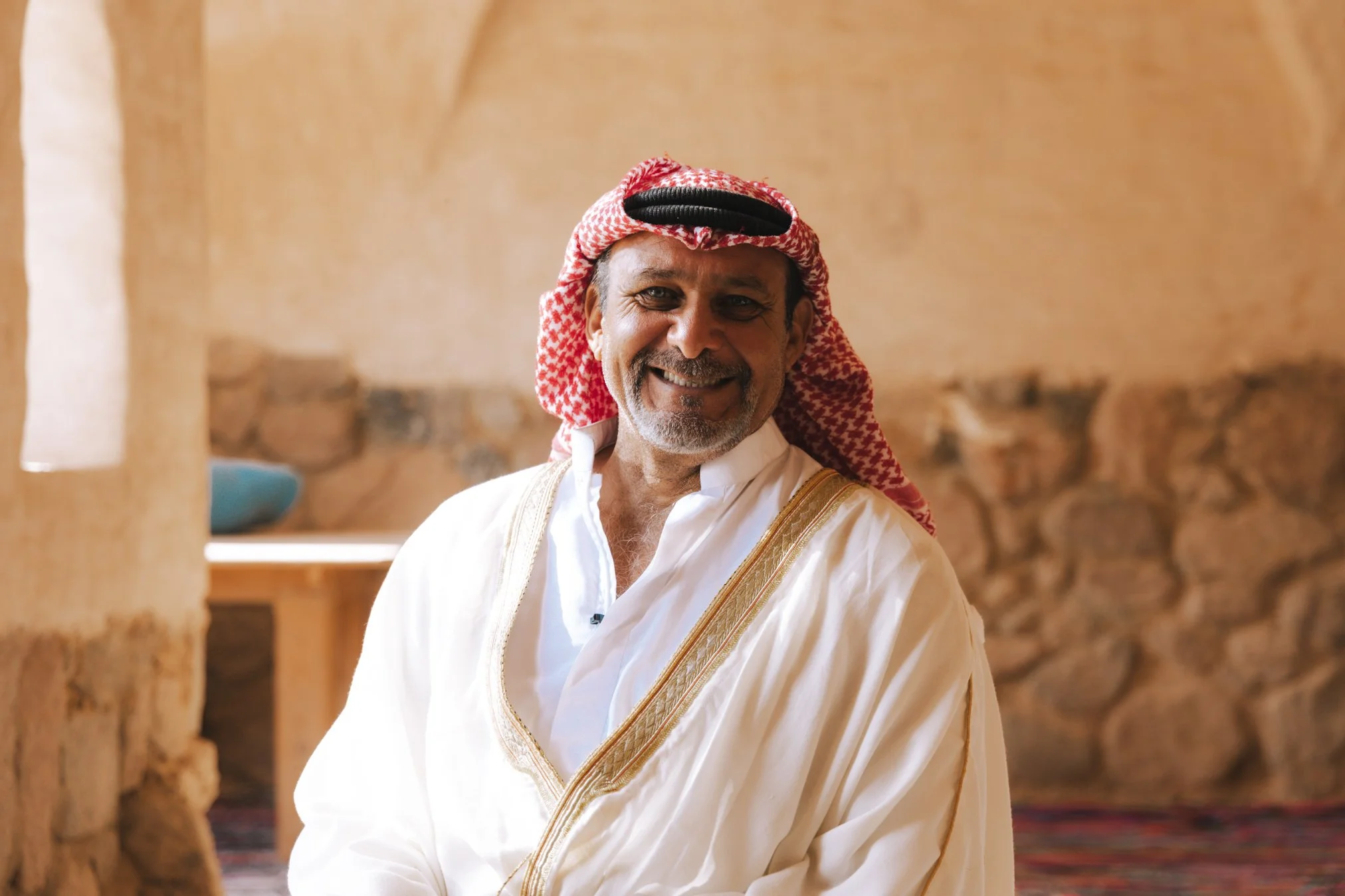 Smiling man wearing traditional Middle Eastern attire, a white robe with gold embroidery, and a red and white checkered keffiyeh headscarf, standing indoors with stone walls in the background.