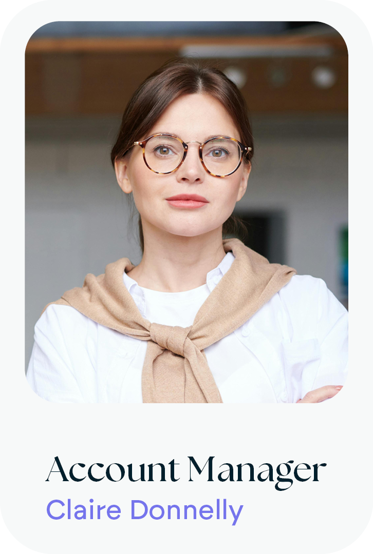 Photograph of a woman with brown hair and glasses, wearing a white shirt and a beige sweater tied around her shoulders, standing in an indoor setting.