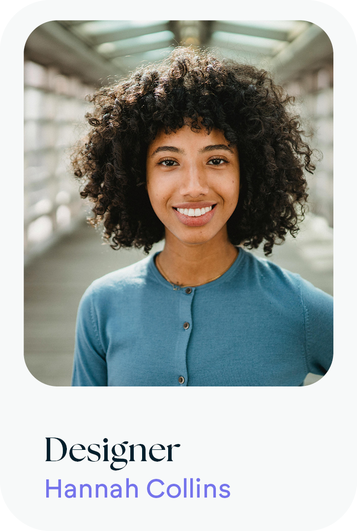 Portrait of a young woman with curly hair smiling, wearing a blue cardigan, standing in a bright, modern indoor space.
