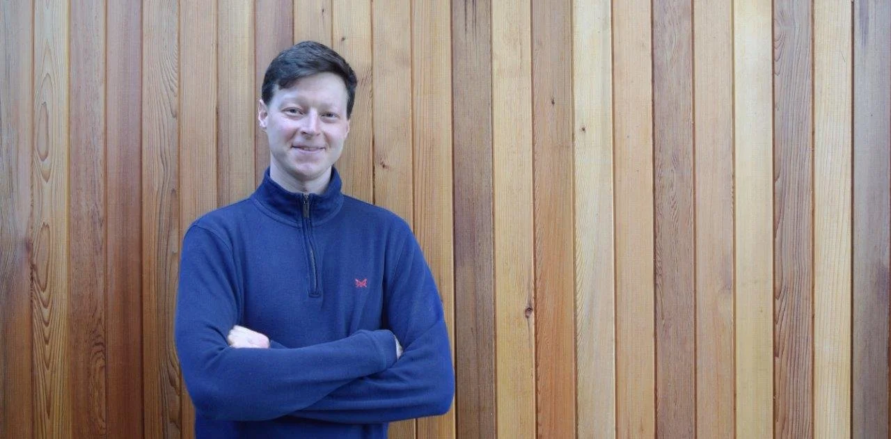 A young man with dark hair, wearing a navy blue zip-up sweater with a small red logo, standing with arms crossed in front of a wooden wall with vertical planks.