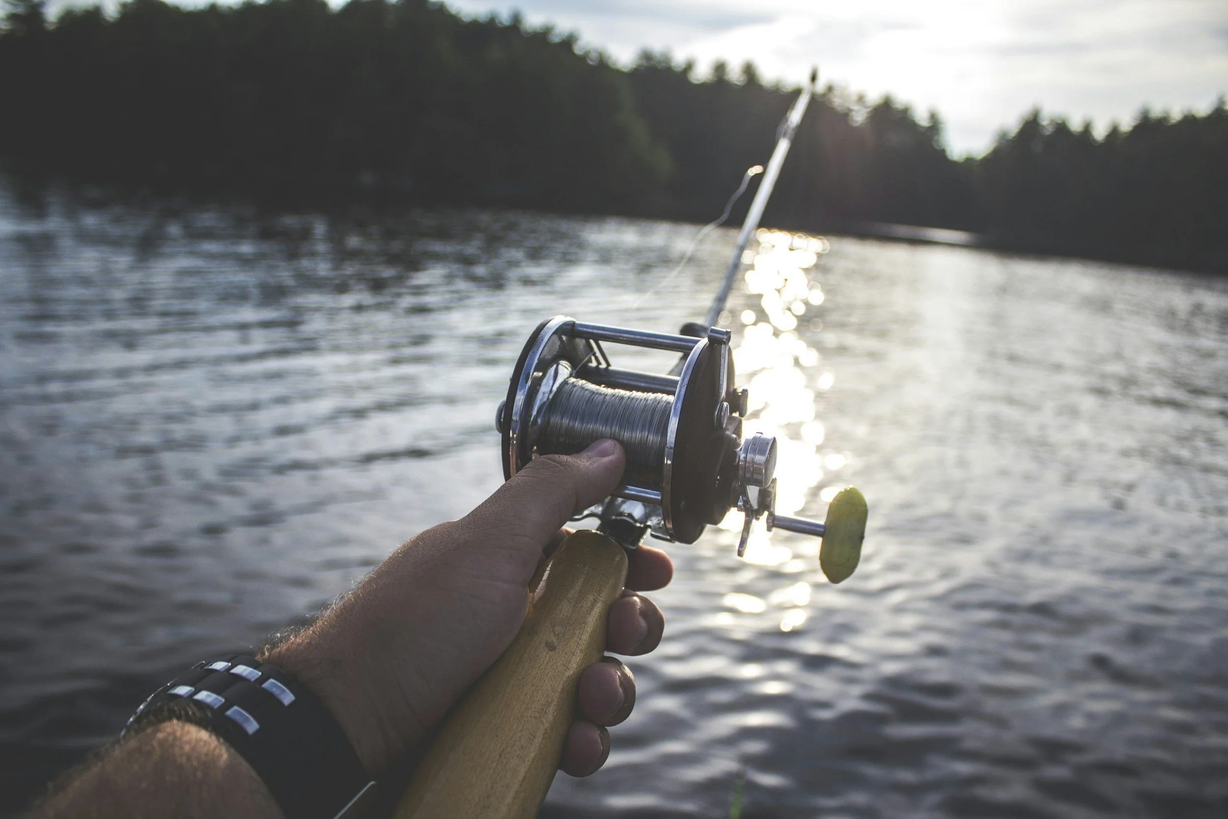 A person's hand holding a fishing rod with a reel, fishing on a body of water during sunset.