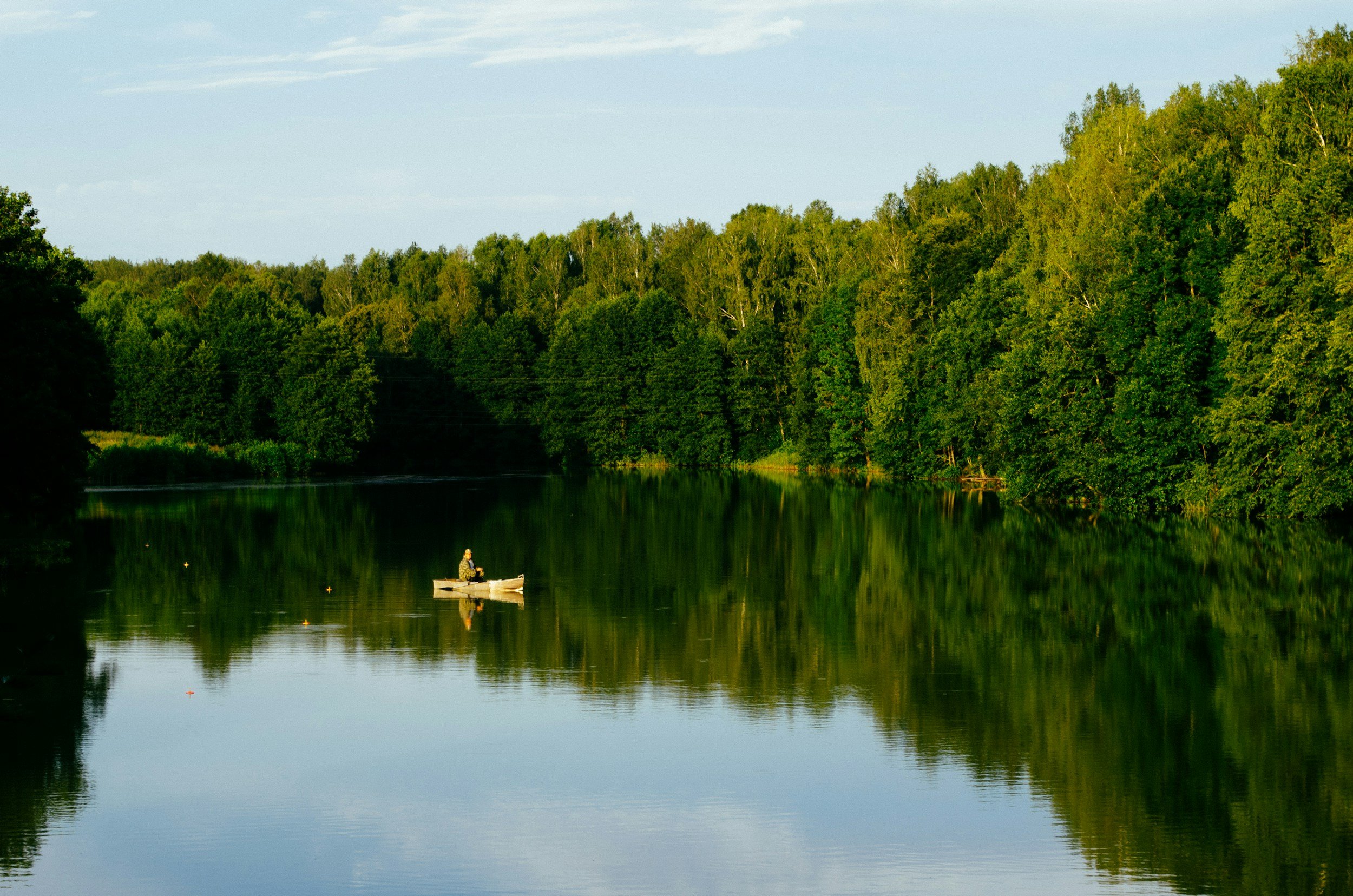 A person fishing from a small boat on a calm river surrounded by dense green trees, with reflections in the water and a partly cloudy sky above.