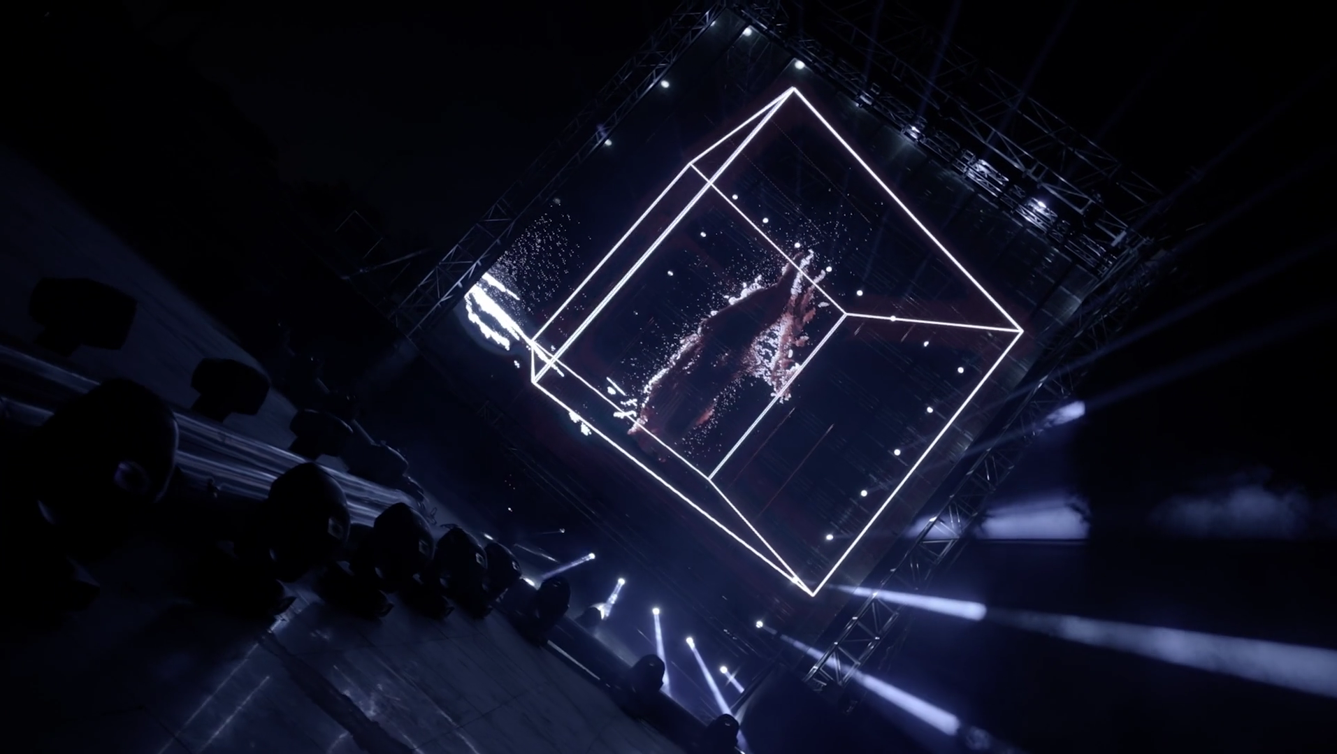 A large outdoor stage with a 3D holographic display of a human figure inside a glowing wireframe cube, surrounded by stage lights and scaffolding, set at night.