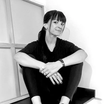 A young woman sitting cross-legged on the floor against a plain wall, smiling slightly, wearing a black long-sleeve shirt and black pants.