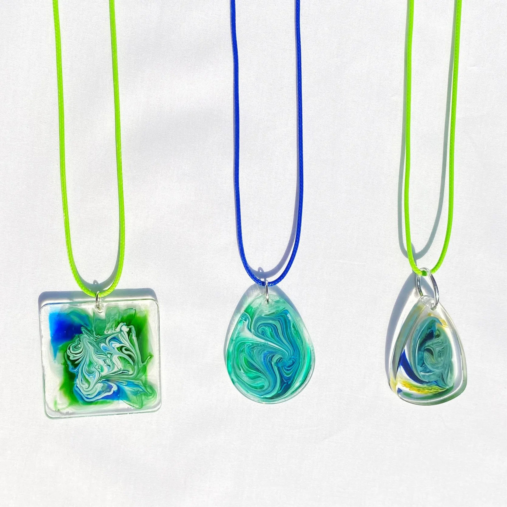 Three resin necklaces with blue, green, and yellow swirled designs hanging from coloured cords on a white background.