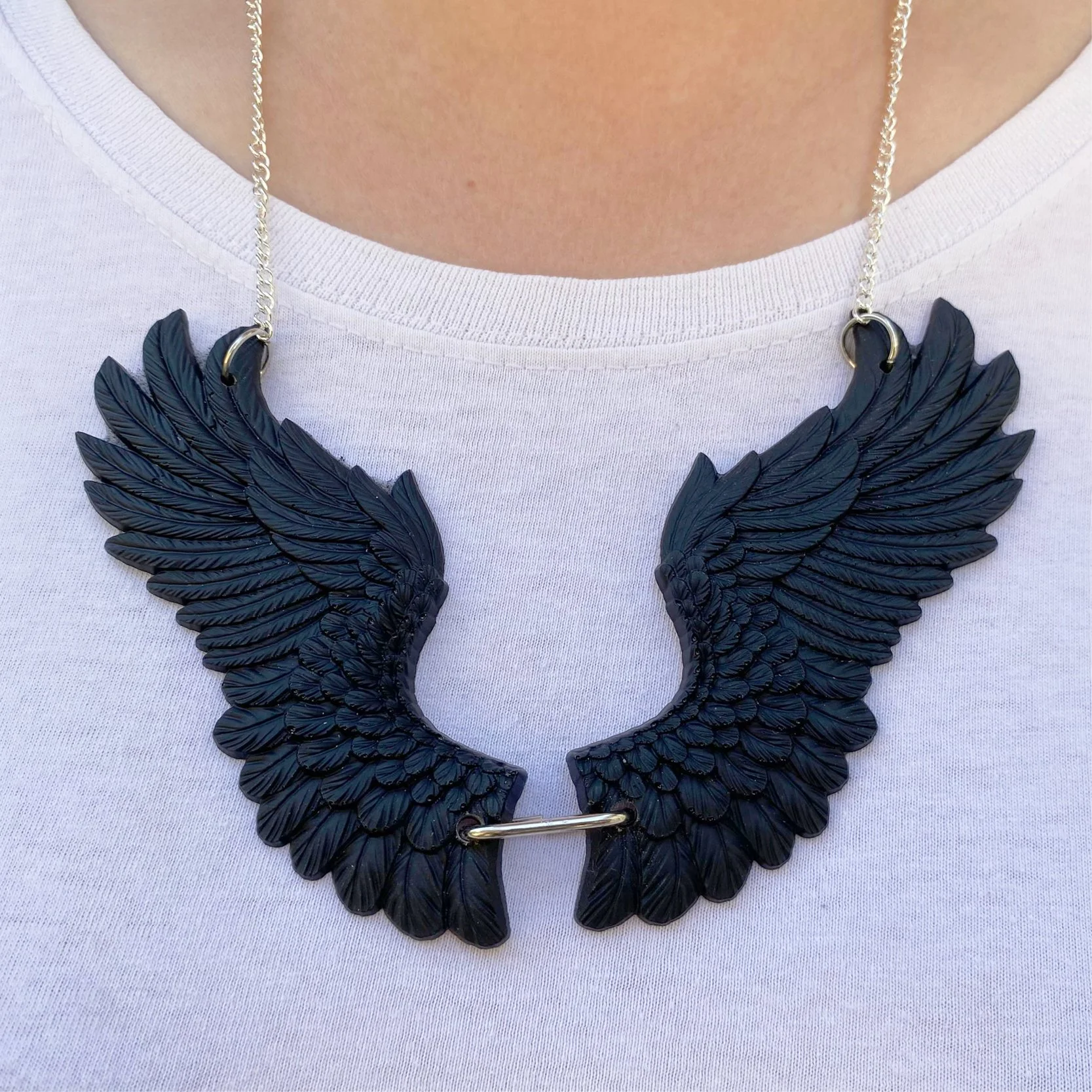 Black wing-shaped necklace with detailed feather design worn around the neck of a person in a white shirt.