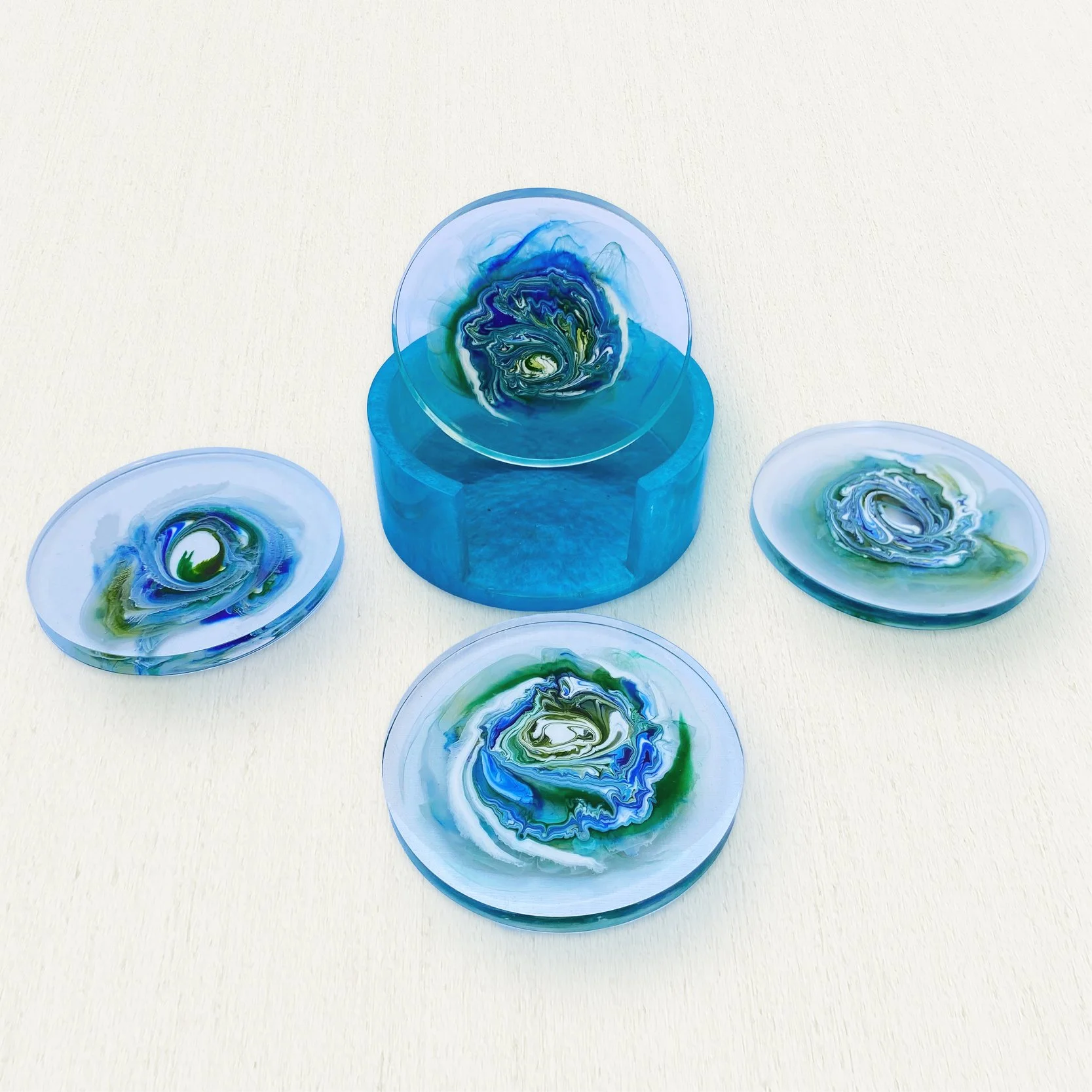 Four resin coasters with abstract blue, green, and white swirling patterns, one elevated on a blue stand.